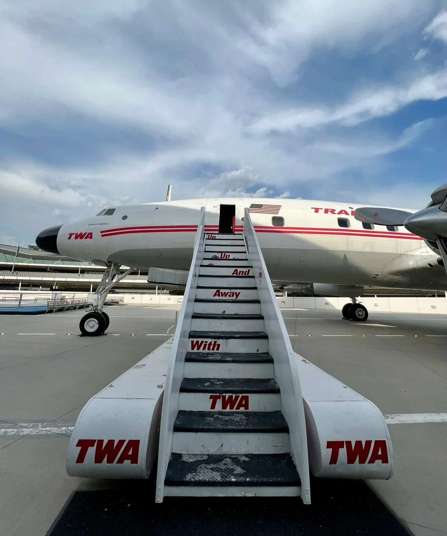 An airplane on the tarmac with a staircase leading up to its door, the staircase has the words "Un Up And Away With TWA" written on it.