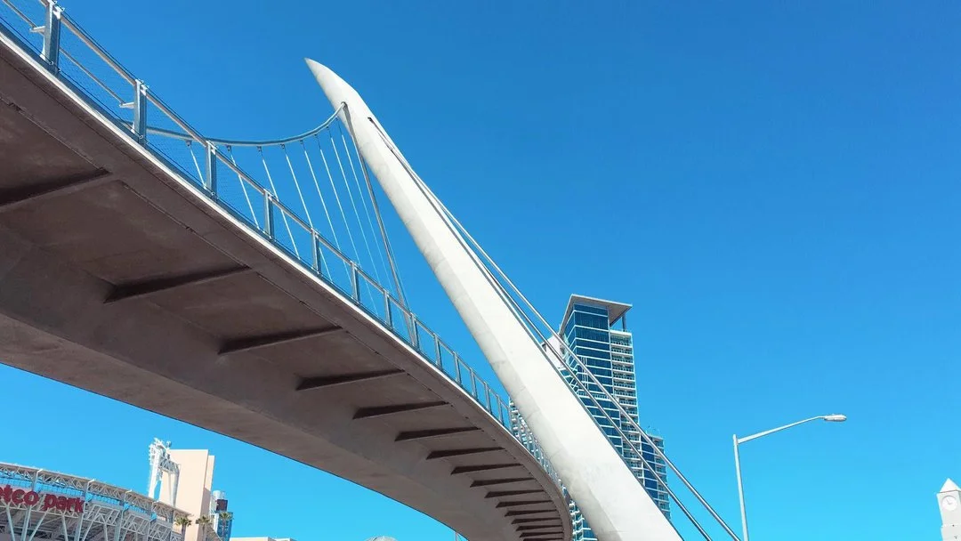 A modern bridge with distinctive white pylon and cable-stayed design, set against a bright blue sky, with tall buildings in the background.