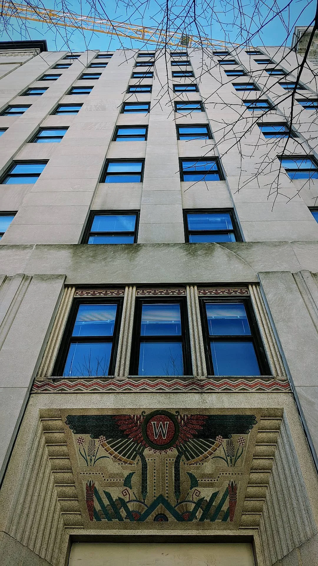 Low-angle view of a tall building with decorative mosaic art featuring a red 'W' surrounded by wings and intricate patterns, set against a clear blue sky with bare tree branches at the top.
