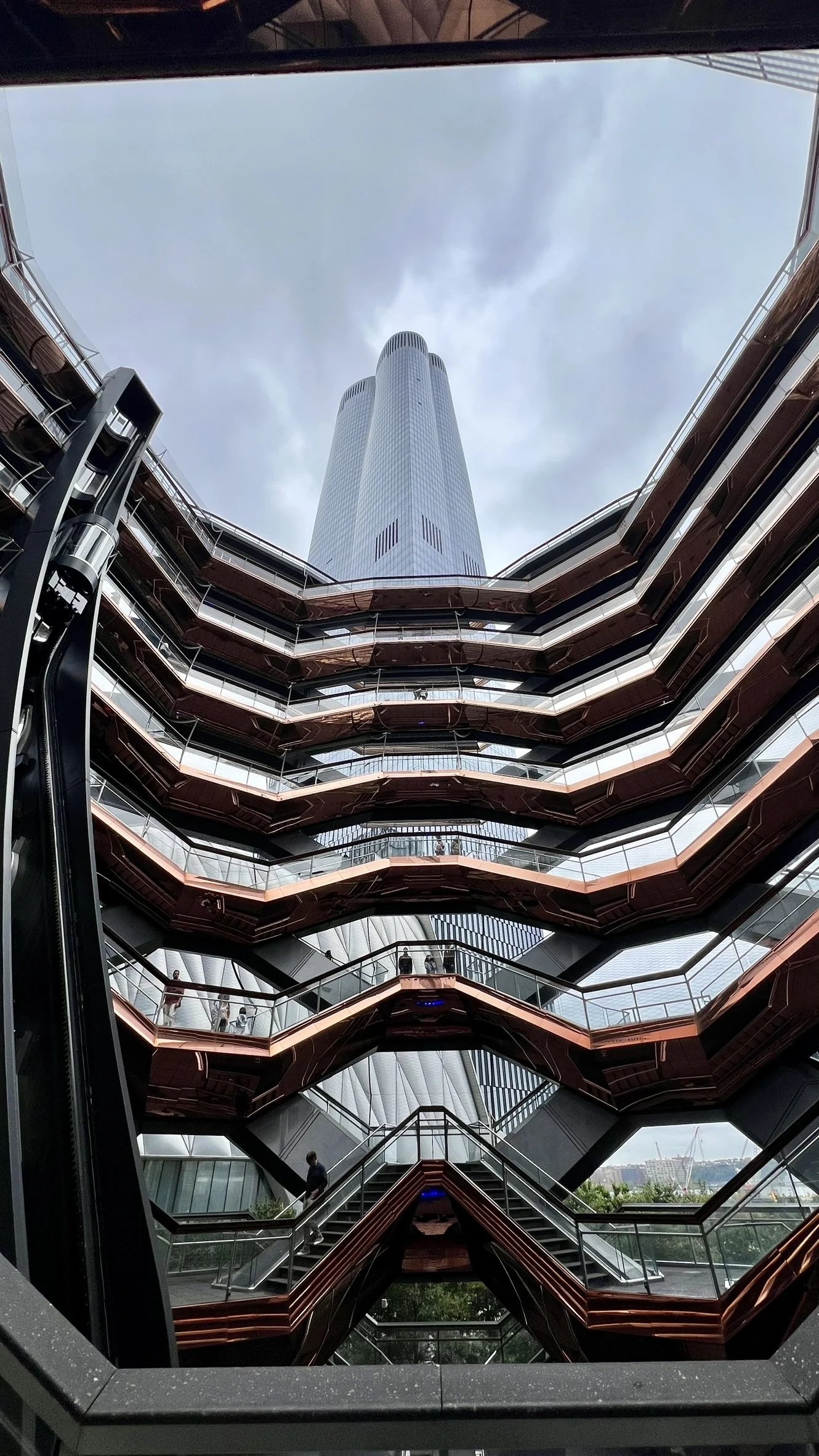 Looking up at the modern, tall building through the glass and metal structure of a multi-level walkway or atrium.