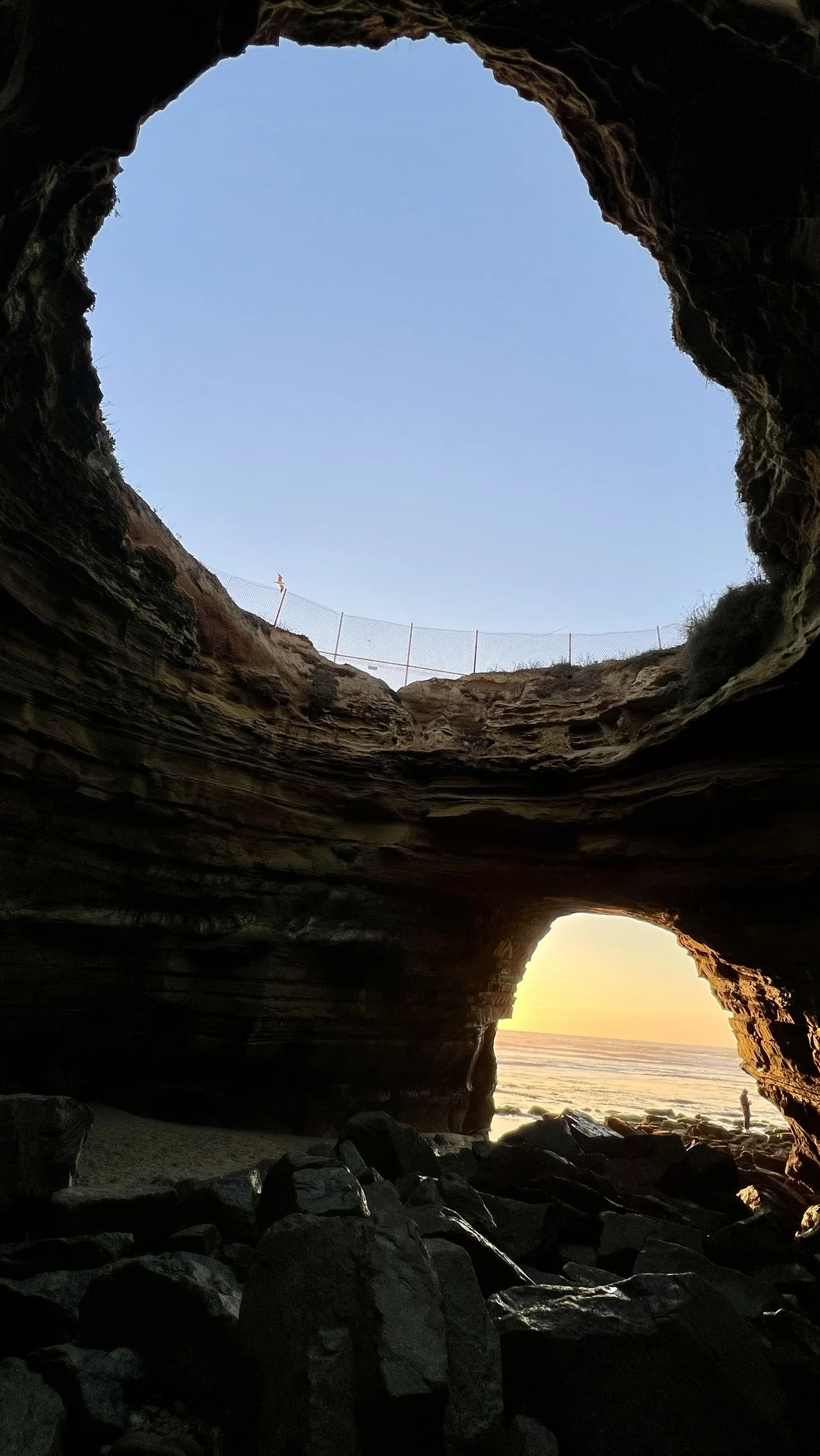 View from inside a sea cave looking out toward the ocean and sunset, with a person standing on the rocks outside.