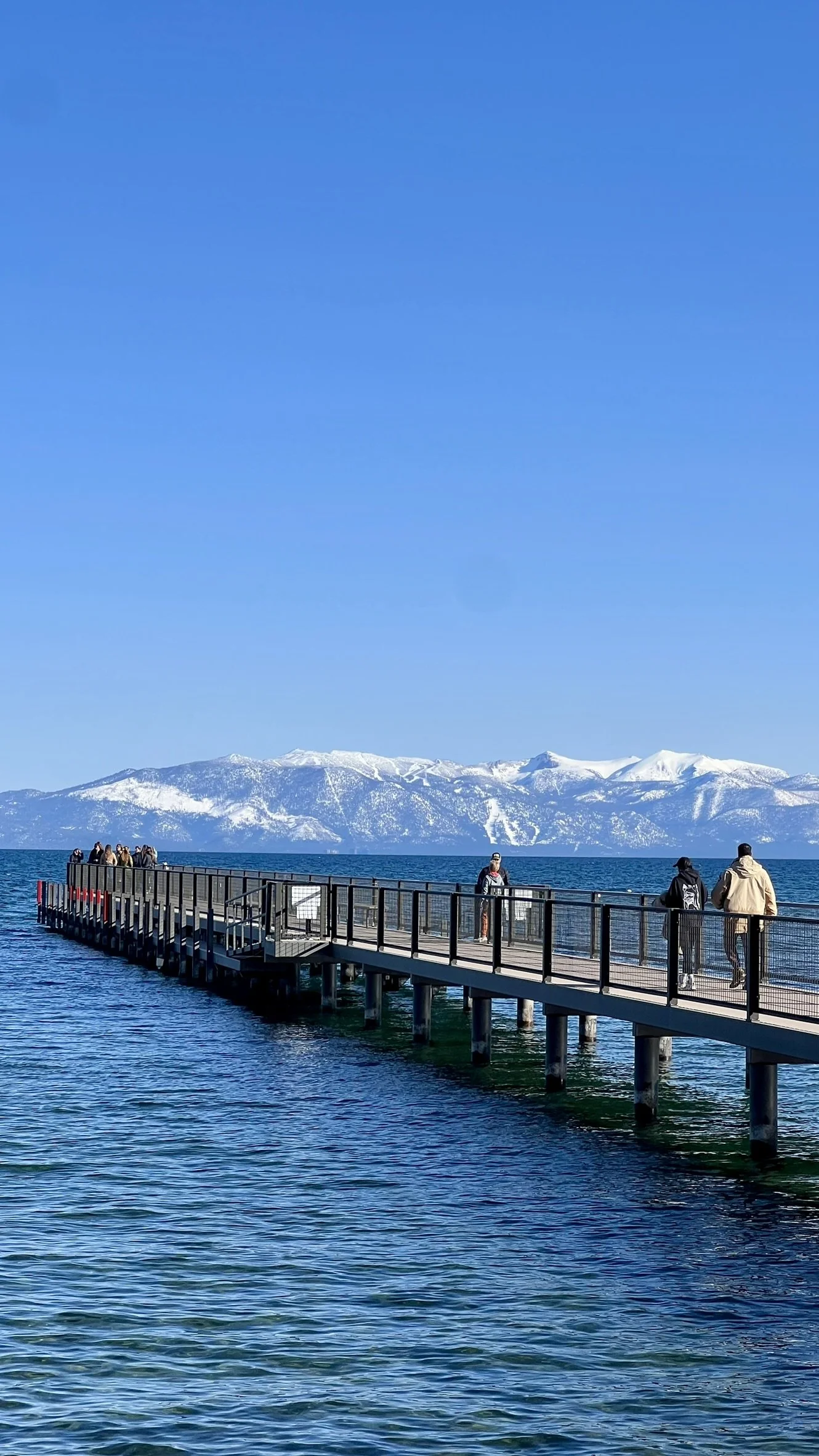 People walking on a pier extending into a large body of water with snow-covered mountains in the background under a clear blue sky.
