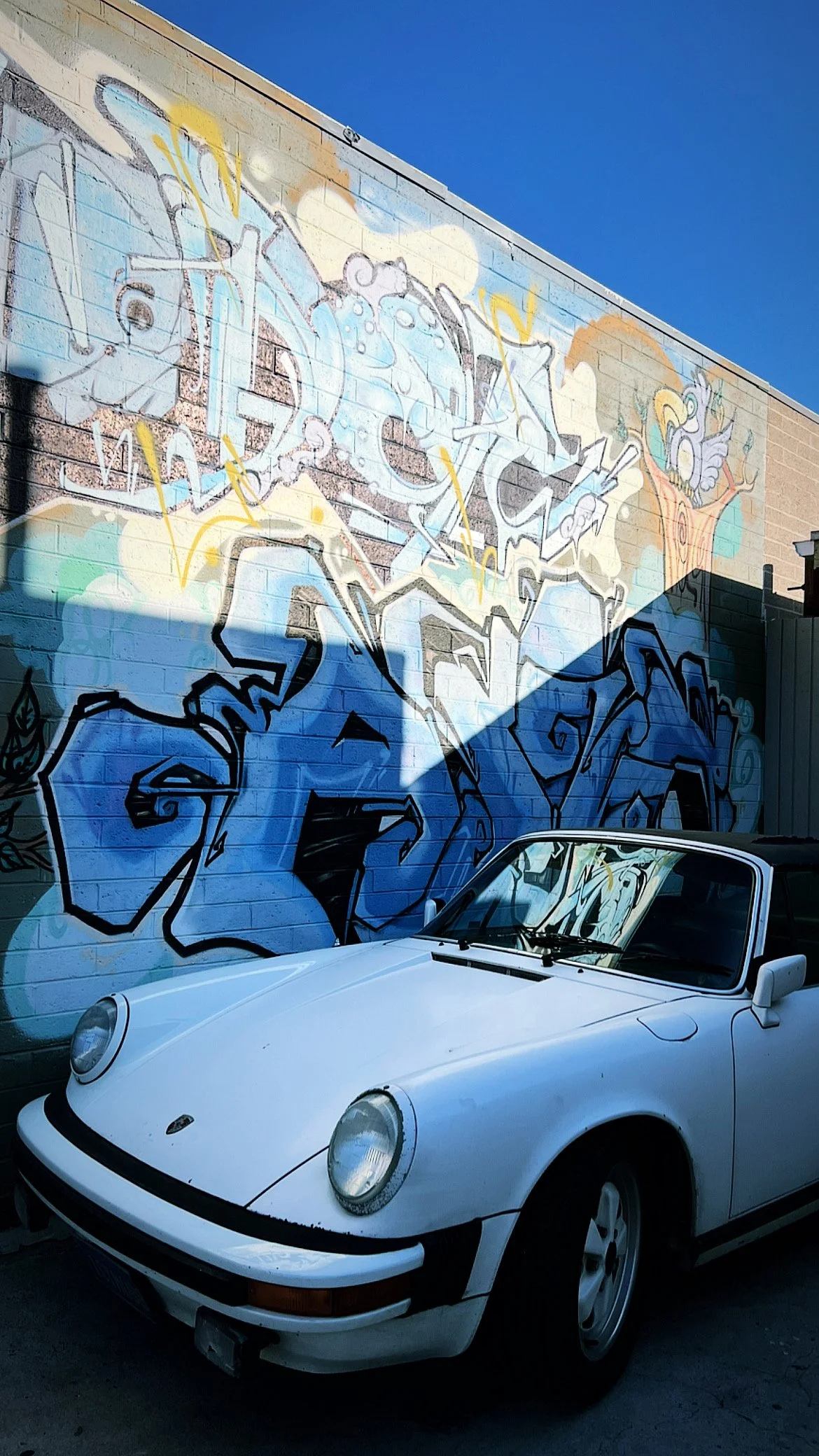 A white vintage Porsche 911 parked in front of a colorful graffiti wall on a sunny day with a clear blue sky.
