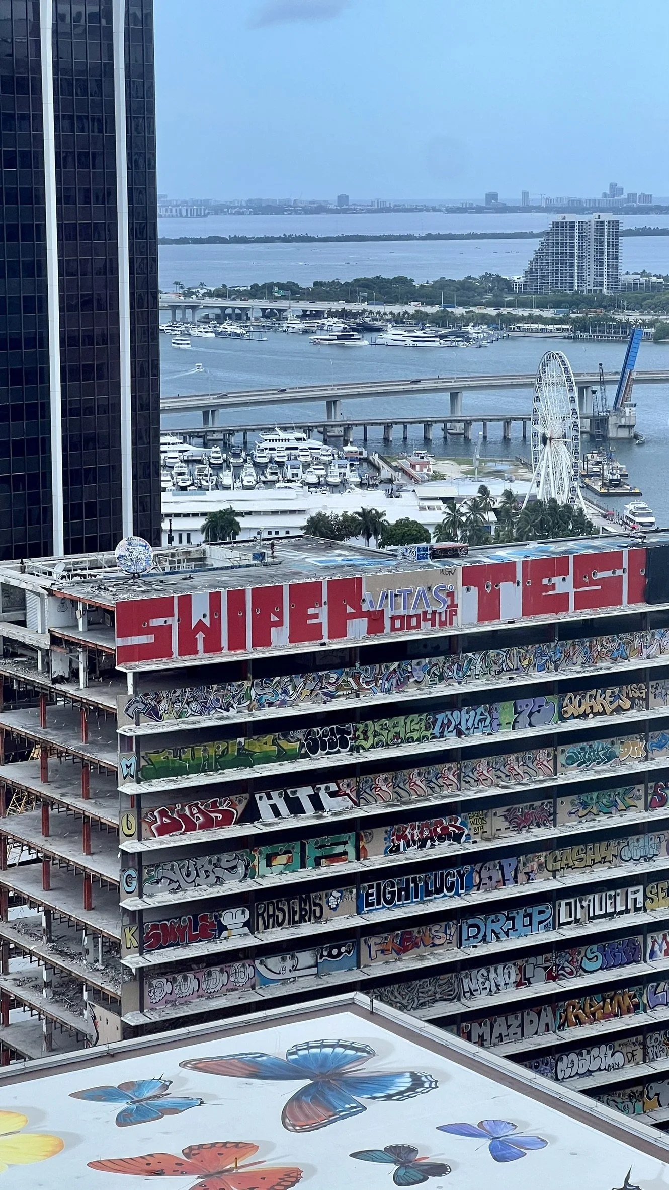 Aerial view of a city landscape with high-rise buildings, a body of water with boats, a Ferris wheel, and an unfinished building covered in colorful graffiti.