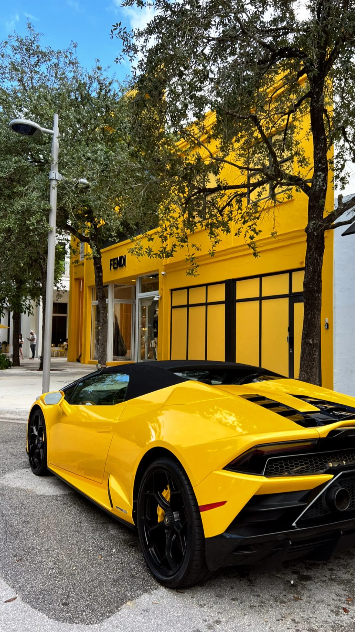 A yellow Lamborghini convertible parked on a city street in front of a yellow building with a store named FELDI. Trees line the sidewalk, and a few pedestrians are seen in the background.