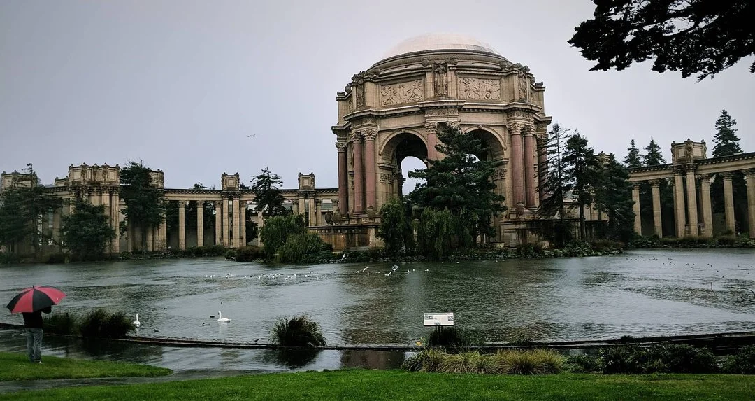 A large classical-style monument with a domed roof and columns, surrounded by water, with trees around and a person holding a pink and black umbrella in the foreground.