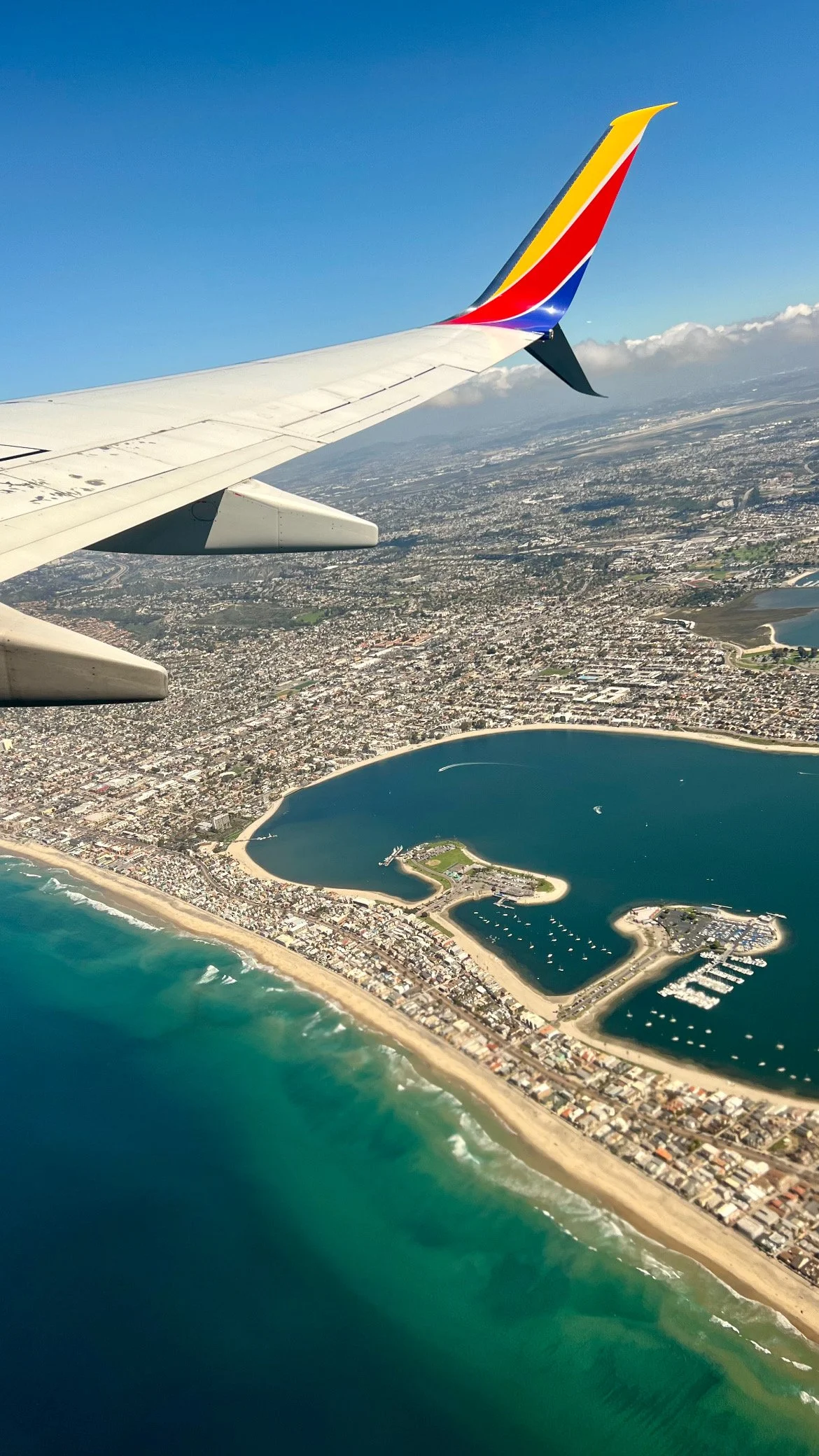 View from airplane window showing coastline, beach, marina, and urban area beneath a clear blue sky.