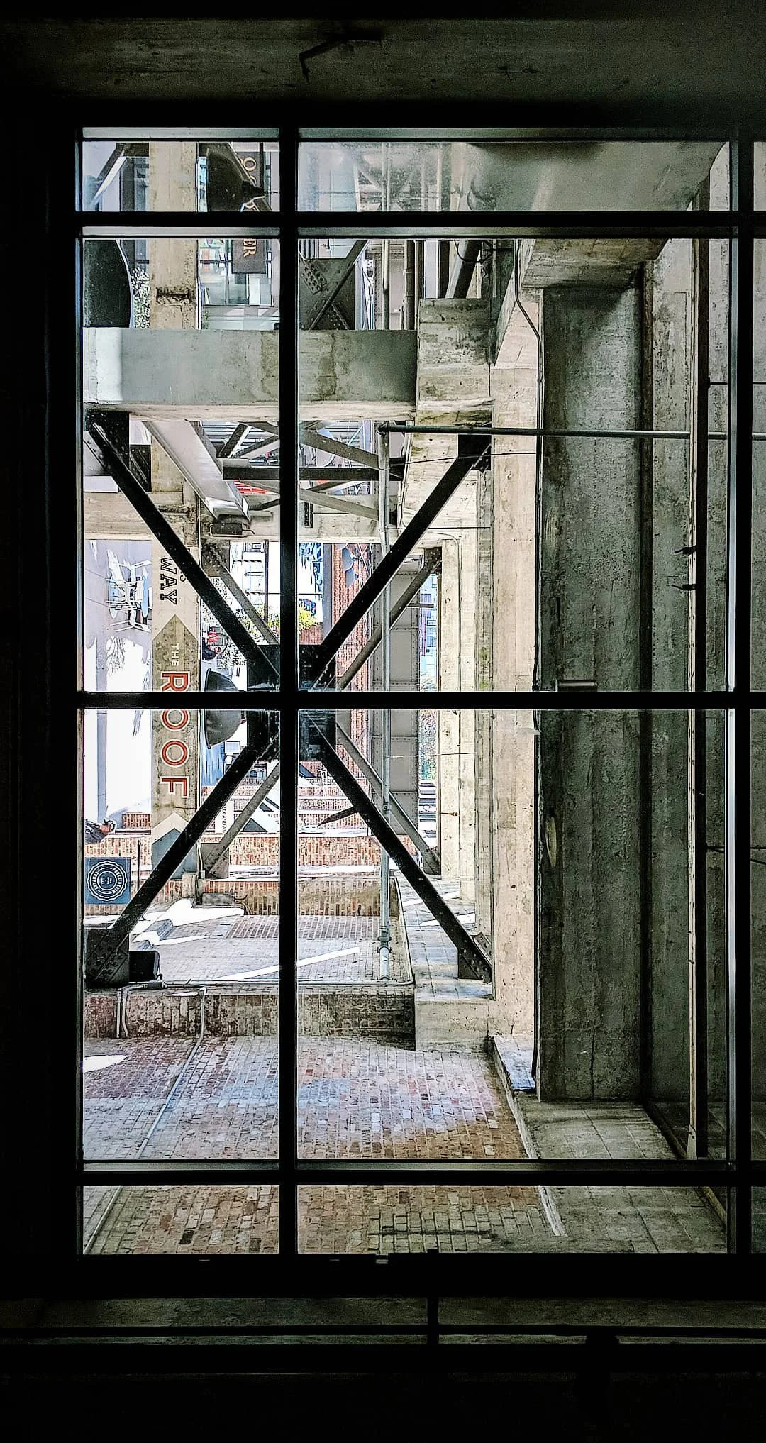 View through a large window of a building under construction, showing exposed brick walls, concrete floors, and metal scaffolding outside.