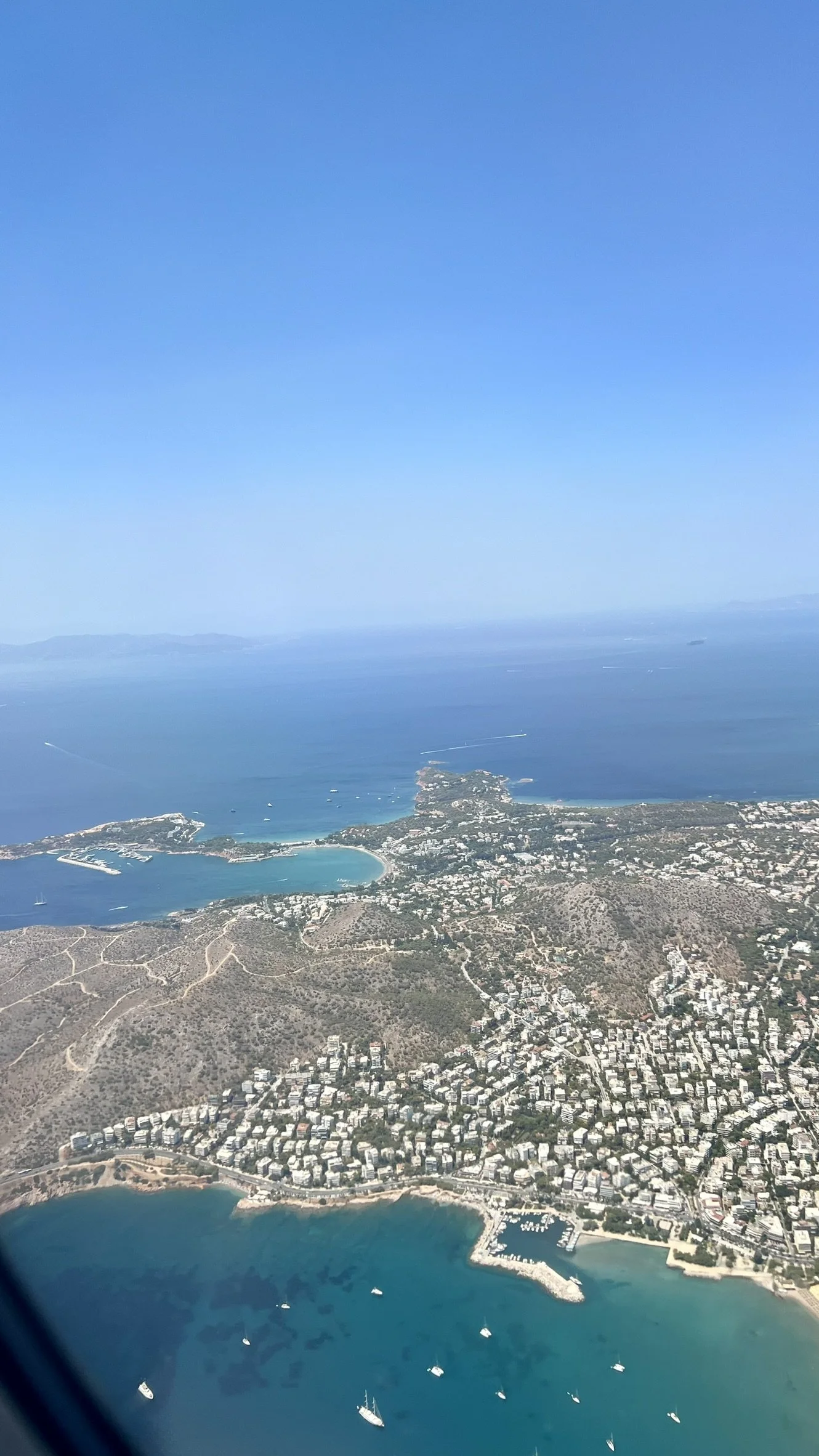 Aerial view of a coastal city with buildings, a marina, and boats in the water, near a large island and open sea under a clear blue sky.