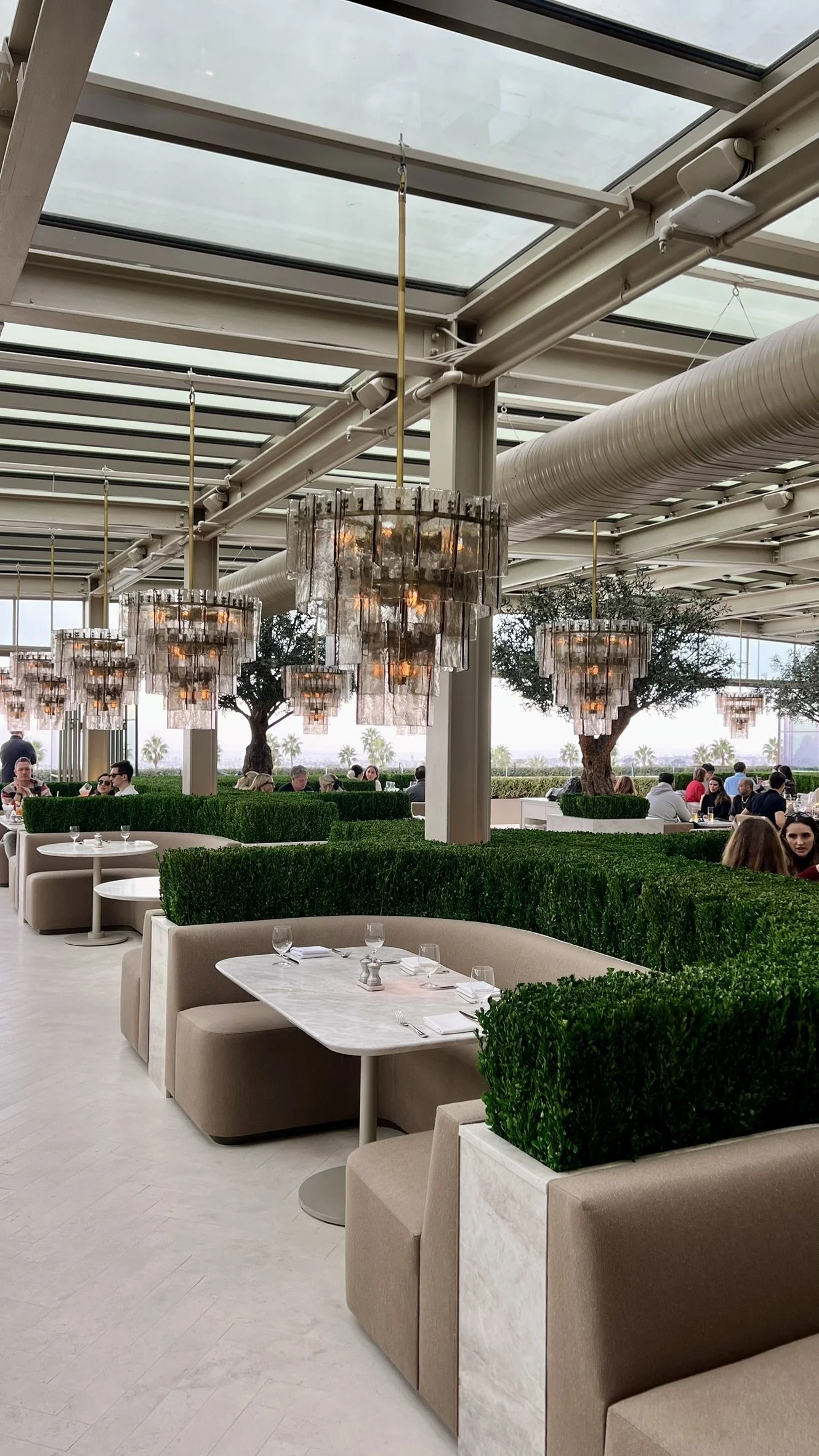 Indoor restaurant with round chandeliers, green hedges, beige seating, and white marble tables set with glassware.