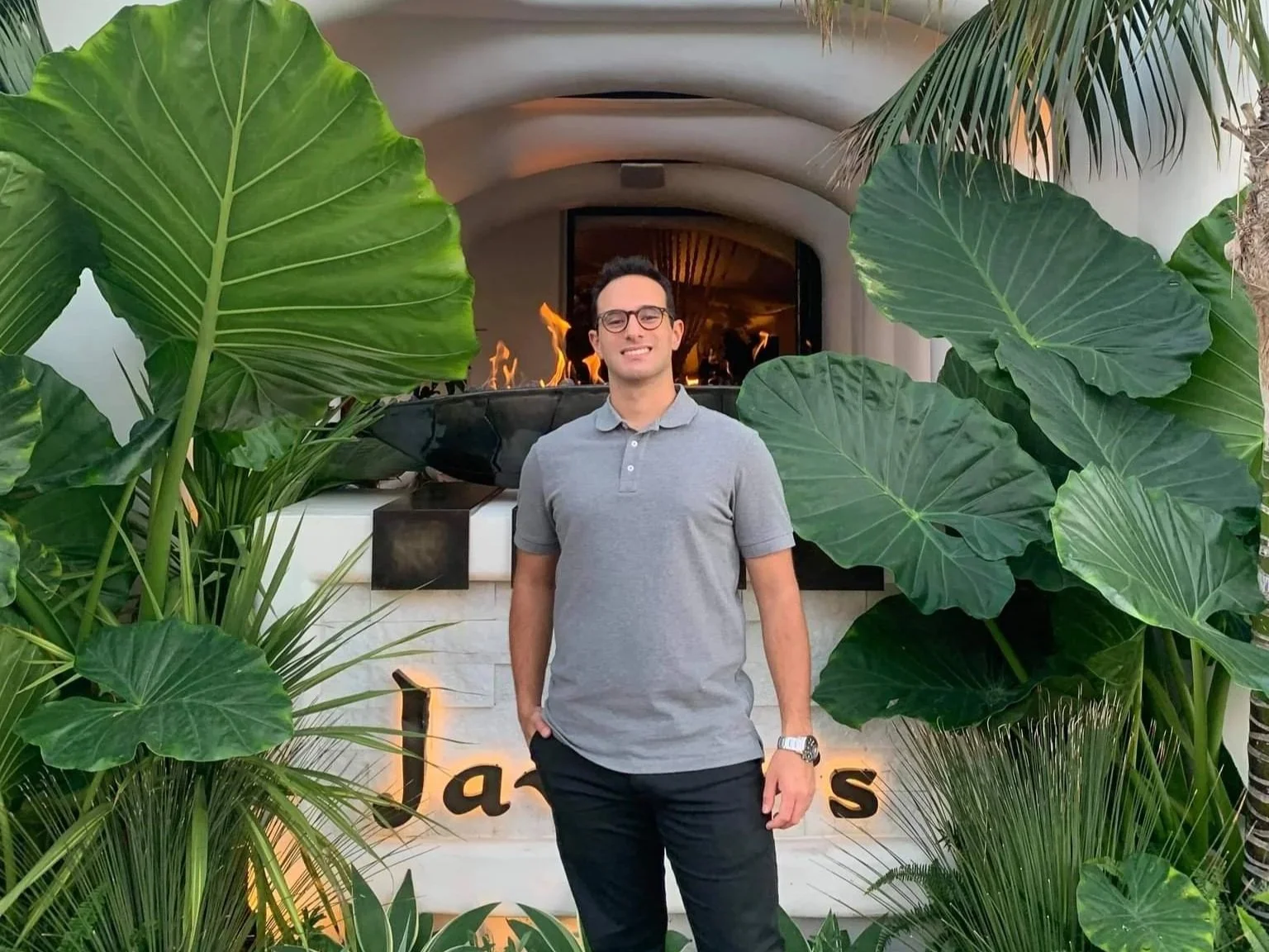 A man standing outdoors in front of a decorative fireplace with flames, surrounded by lush green tropical plants, smiling and wearing glasses, a gray polo shirt, and dark pants.