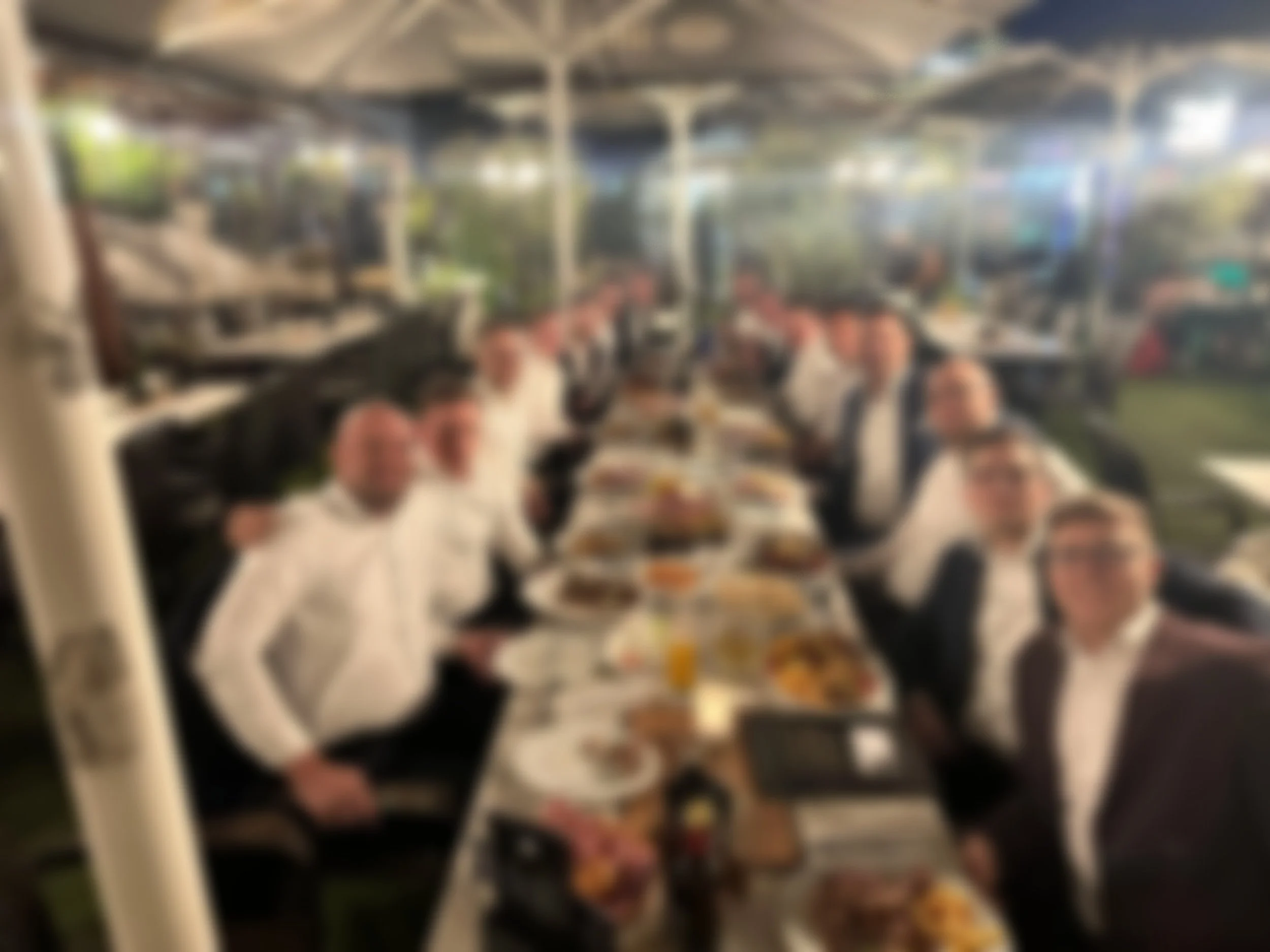 Eine Gruppe von Menschen in formeller Kleidung sitzt an einem langen gedeckten Tisch bei einem Essen in einem Restaurant im Freien, welche das Team von SZ Finanz darstellt.