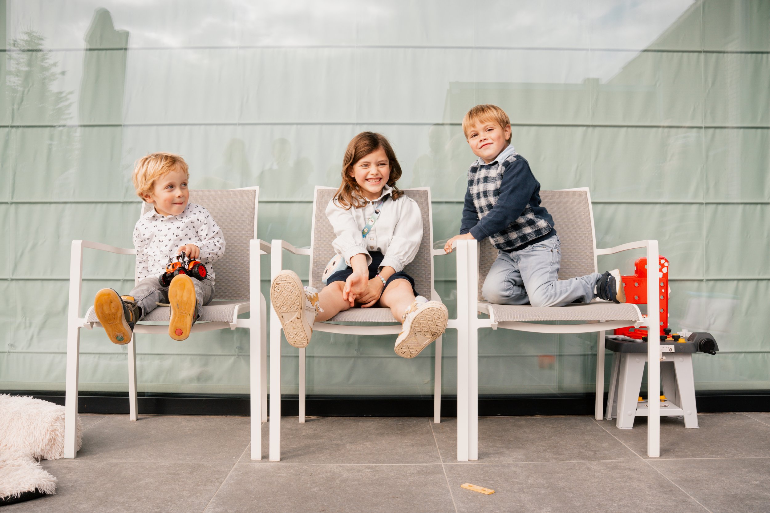 Drie jonge kinderen zitten en hangen op witte stoelen in een ruimte met een groene achtergrond, lachend en spelend met speelgoed.