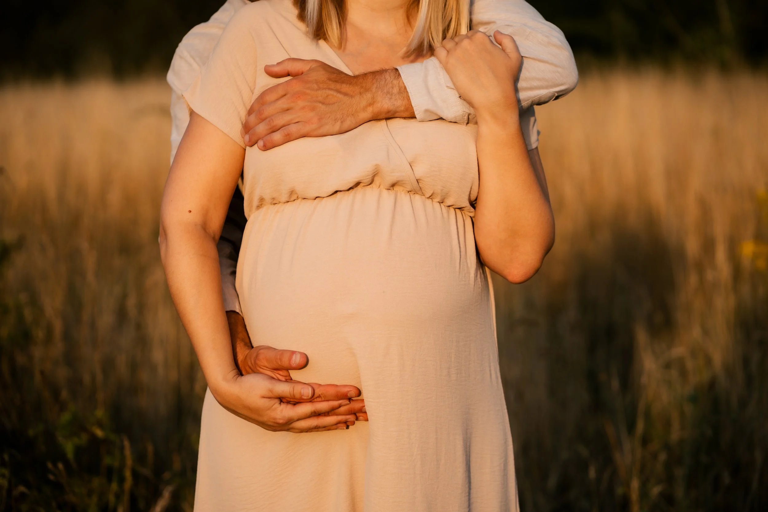 Een zwangere vrouw wordt omringd door twee mensen, met hun handen op haar buik en schouders, tijdens zonsondergang in een veld.