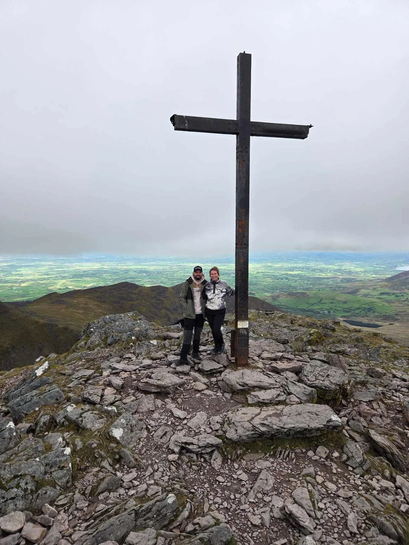 Twee mensen staan naast een groot metalen kruis op een bergtop, met een panoramisch uitzicht op het platteland en de heuvels onder een bewolkte hemel.