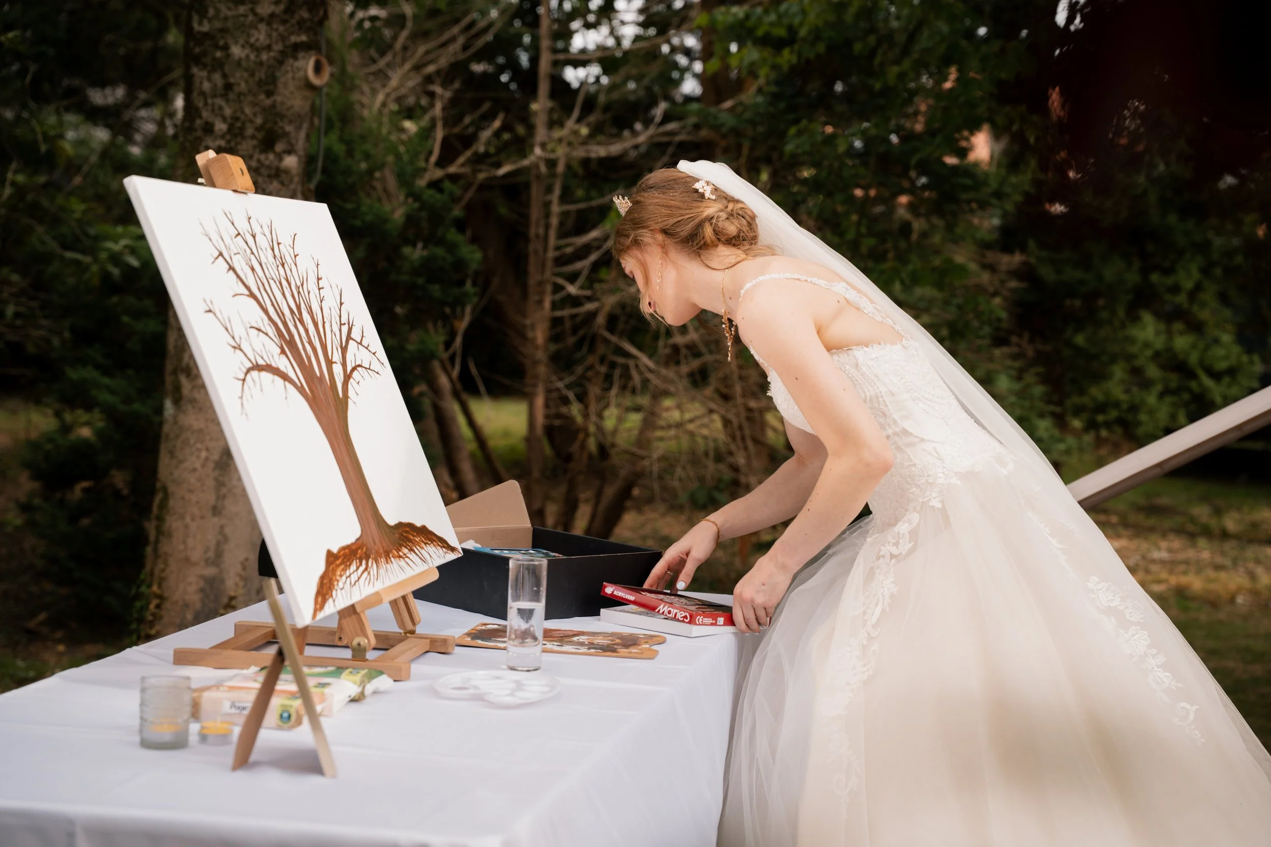Bruidsmeisje in witte trouwjurk die op een tafel staat en schildert op een doek met een boom erop