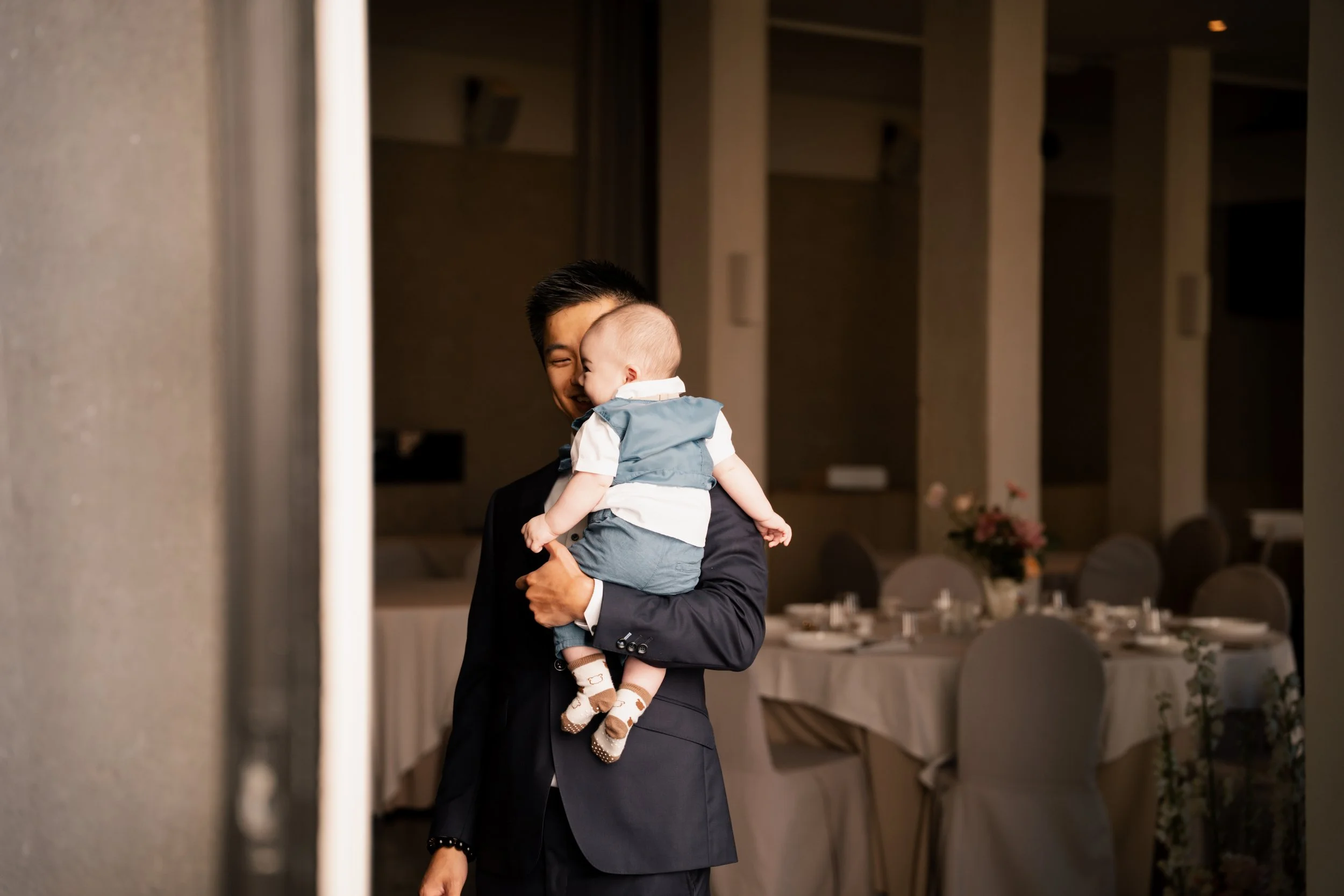 Man in pak op een feestzaal die een baby vast heeft. De zaal is mooi ingericht met gedekte tafels en bloemen.