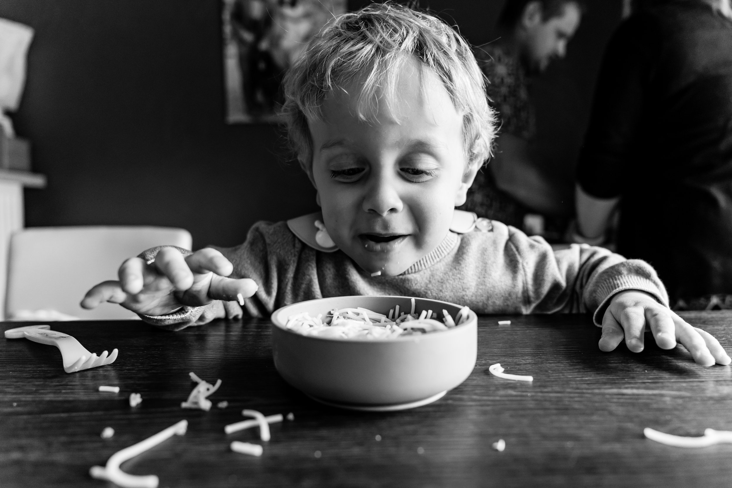 Een jonge jongen met blond haar kijkt verbaasd naar een kom spaghetti op tafel, met pastaresten rondom.