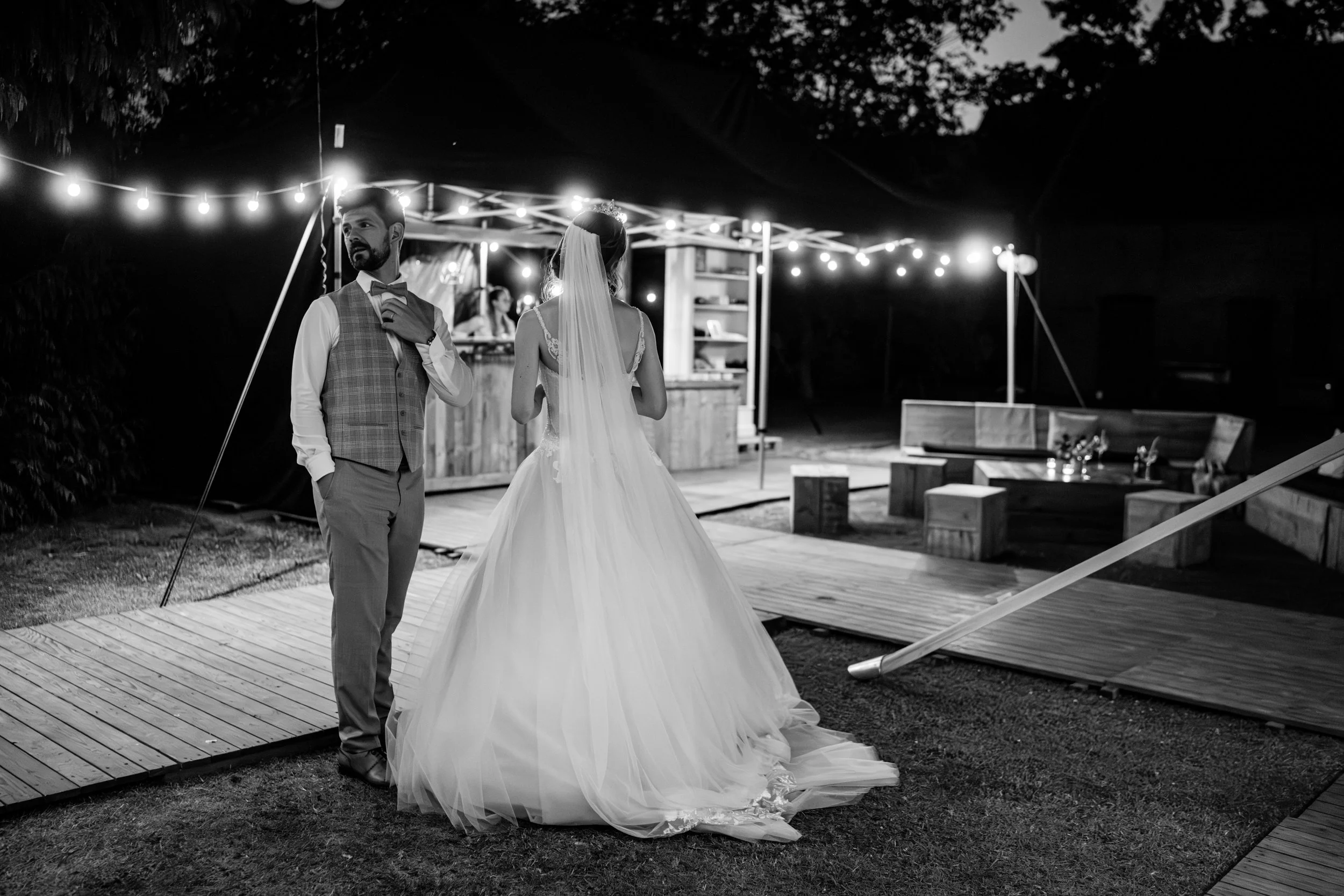 Een bruid en een bruidegom staan buiten onder tuinverlichting tijdens een avond bruiloft, met een bar en zitgedeelte op de achtergrond.