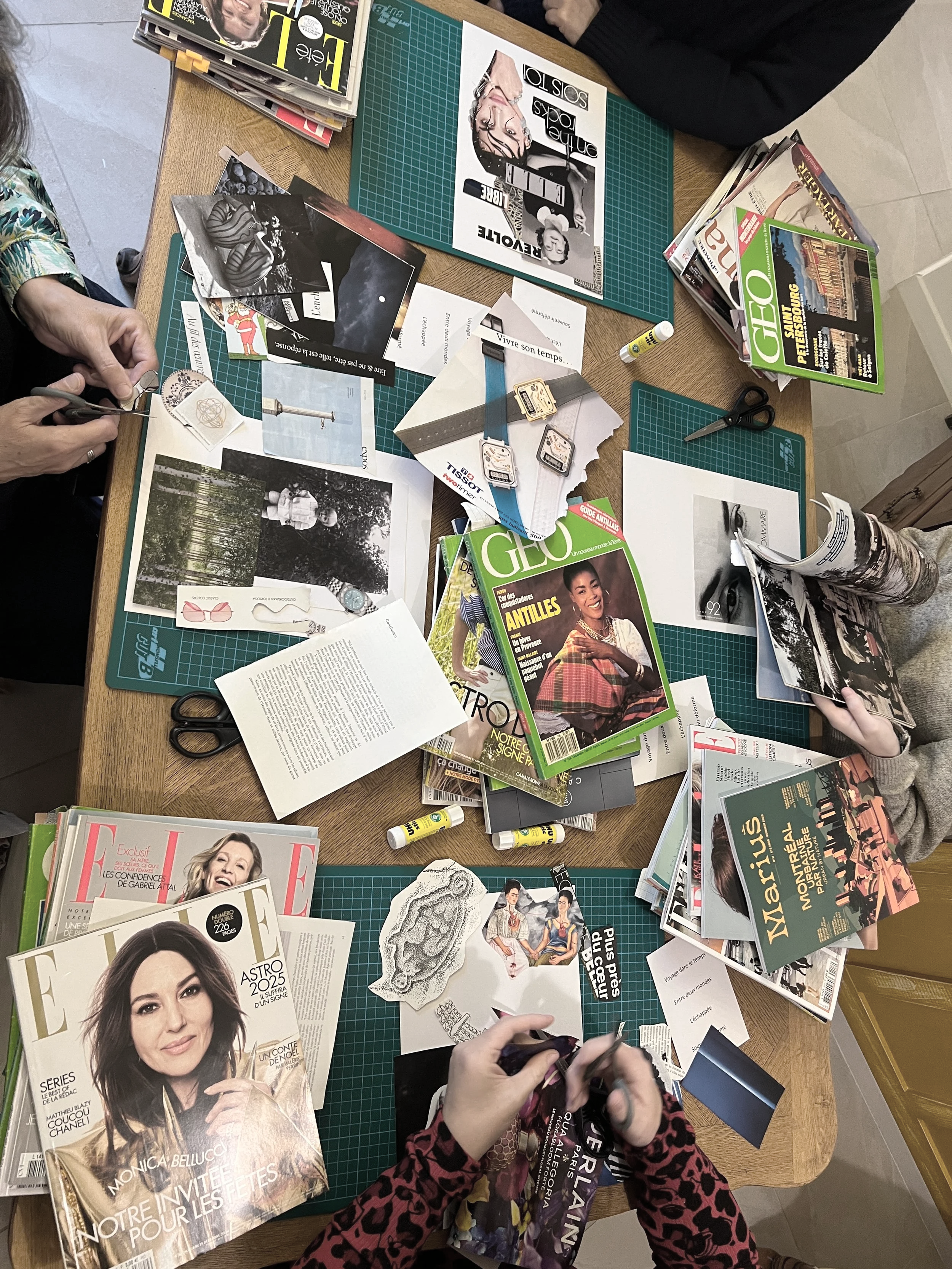 A cluttered table with magazines, printed photos, scissors, glue sticks, and paper cutouts, suggesting an arts and crafts or collage activity involving magazines and photographs.
