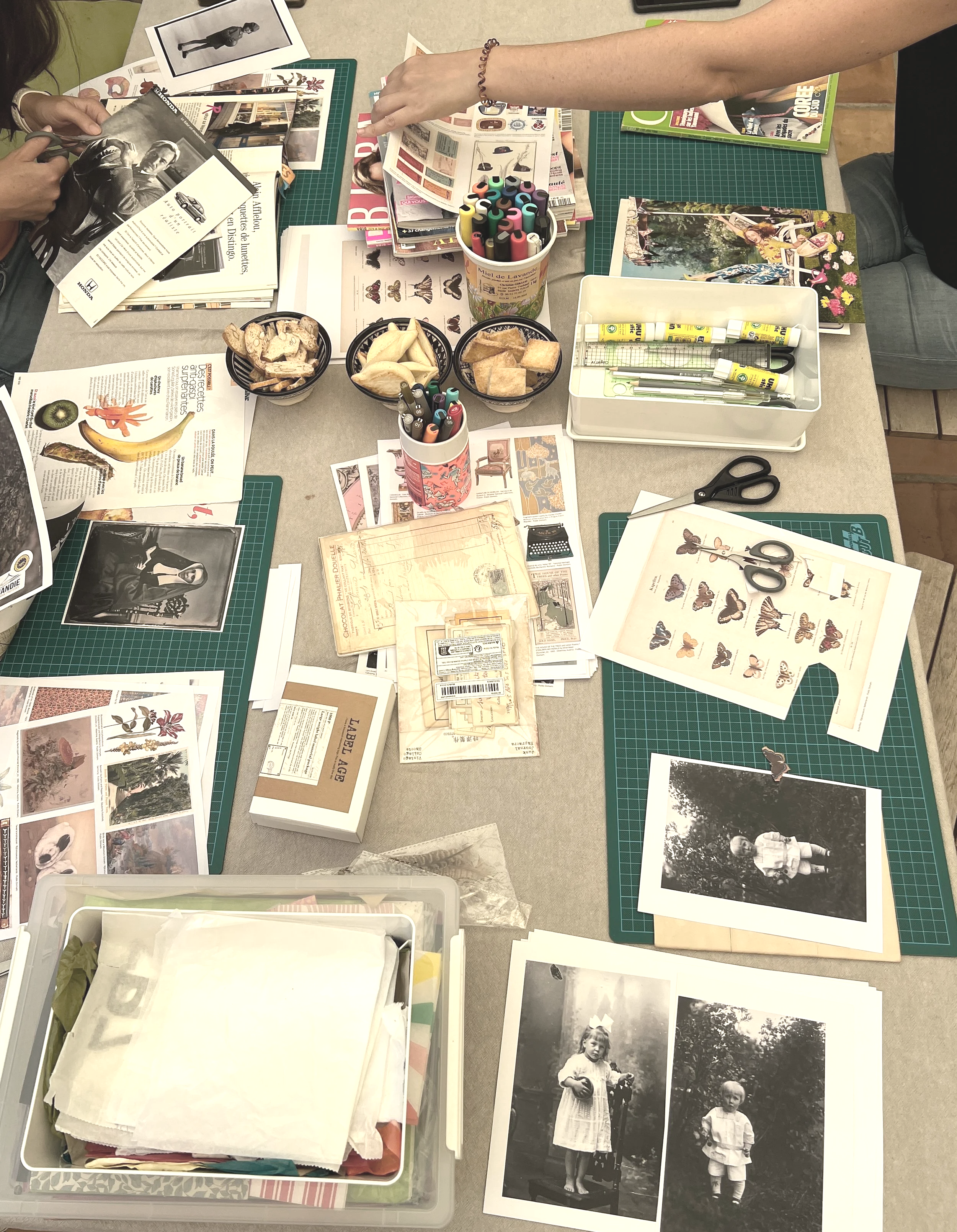 A cluttered table with magazines, photographs, scissors, markers, and various papers, possibly for scrapbooking or craft activities.