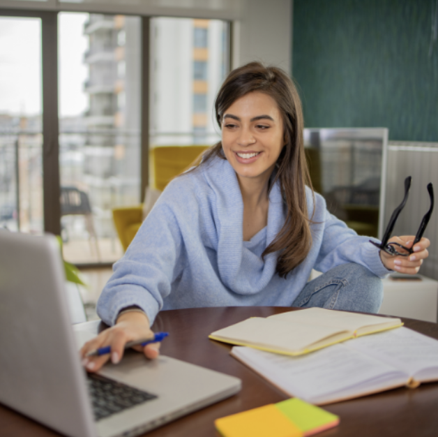 Young woman sitting at a desk with open books, a laptop, and colorful sticky notes, smiling and holding a pair of glasses in a bright, modern office.