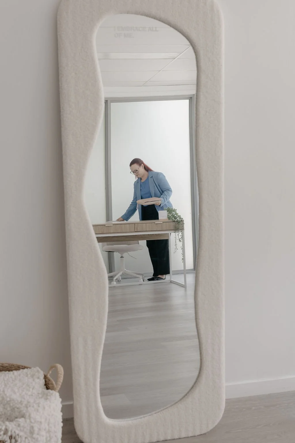 A woman with red hair and glasses, dressed in a blue blazer and black pants, looking at documents at a desk in an office space, reflected in a decorative full-length mirror with a soft, textured frame.