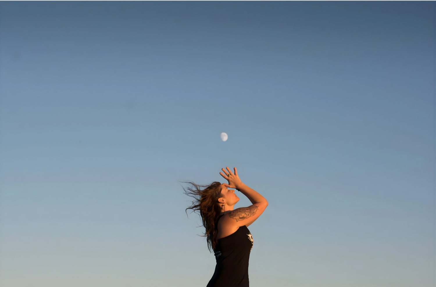 A woman with flowing hair standing outdoors under a blue sky, catching the moon with her hand.
