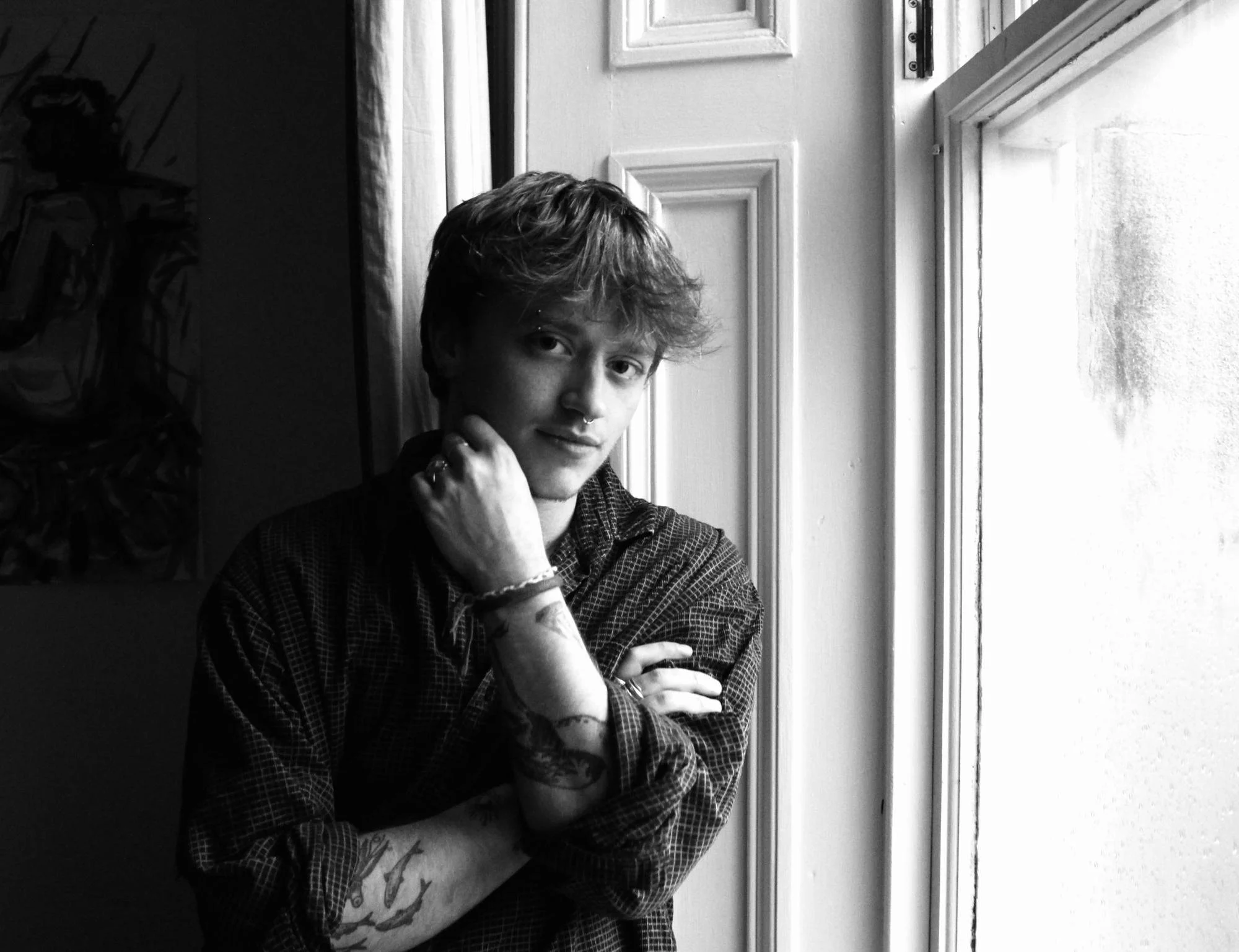 A young man with wavy hair and tattoos on his arms stands near a window, looking at the camera with a slight smile, in black and white.