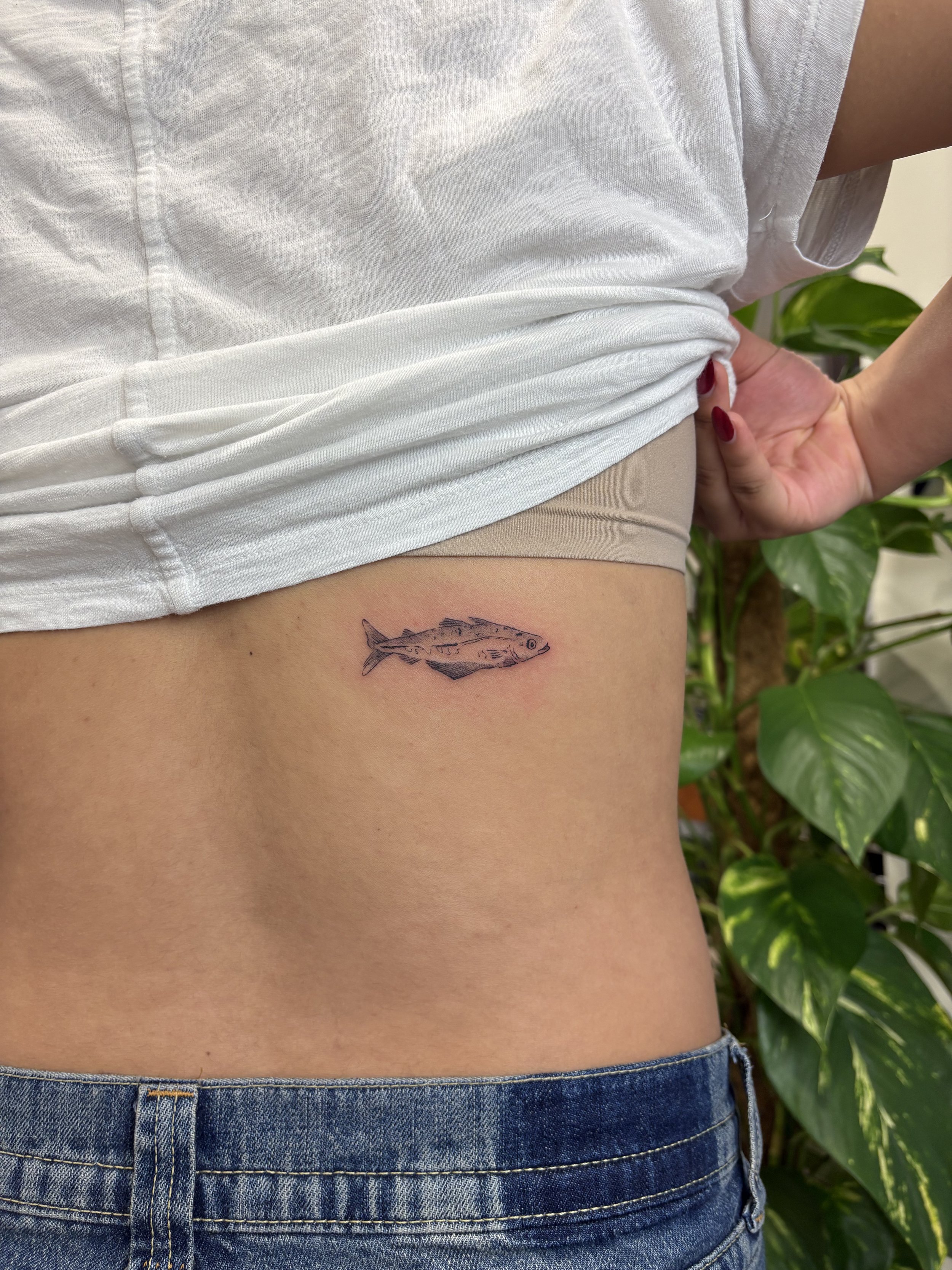 Close-up of a person's side with a fish tattoo on the lower ribcage, wearing a white shirt and blue jeans, with a background of green leafy plants.