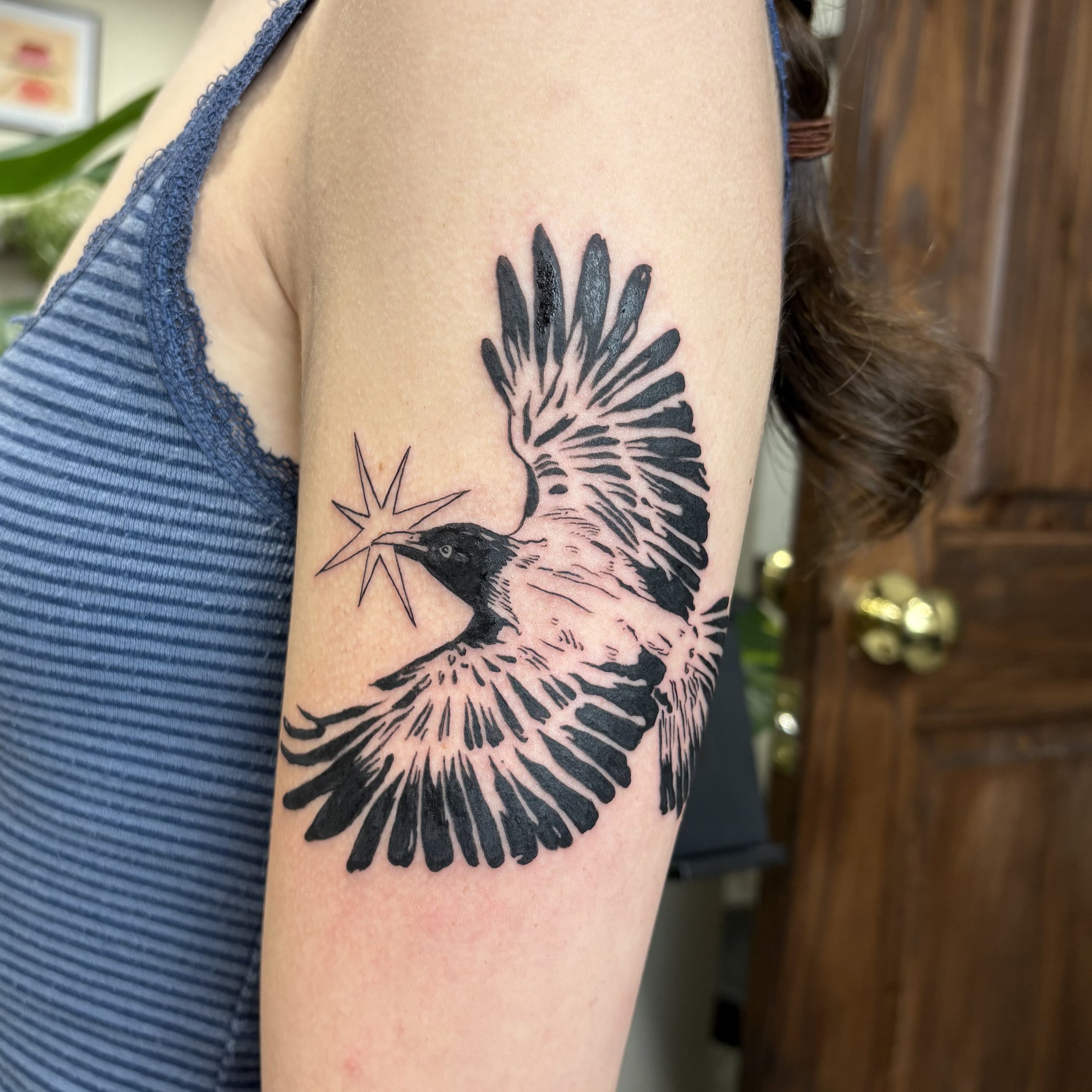 A tattoo of a bird, possibly a woodpecker, with black ink, holding a star-shaped object in its beak, on the person's upper arm.