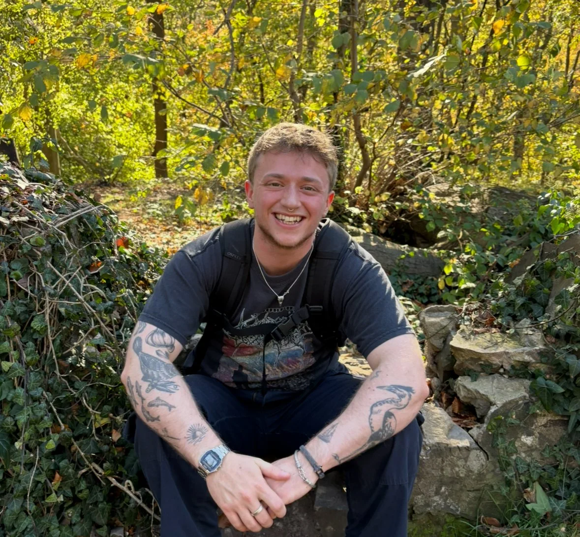 A young man with tattoos on his arms, wearing a black t-shirt, sitting outdoors on a rock. He is smiling, with a backpack, surrounded by green foliage in a daytime setting.