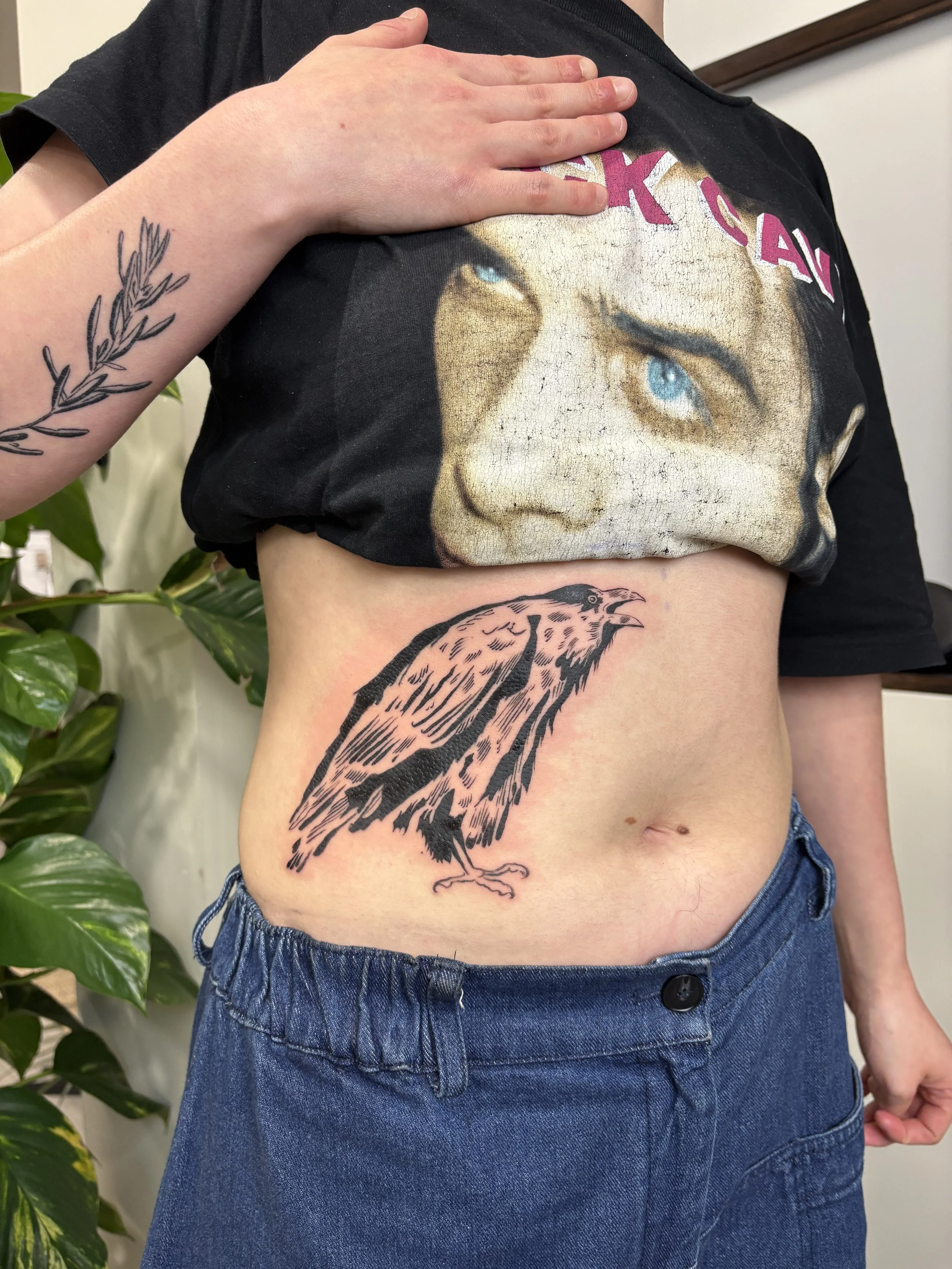 Person with black T-shirt lifting it to reveal a tattoo of a bird on their side and a tattoo of a face on their chest.
