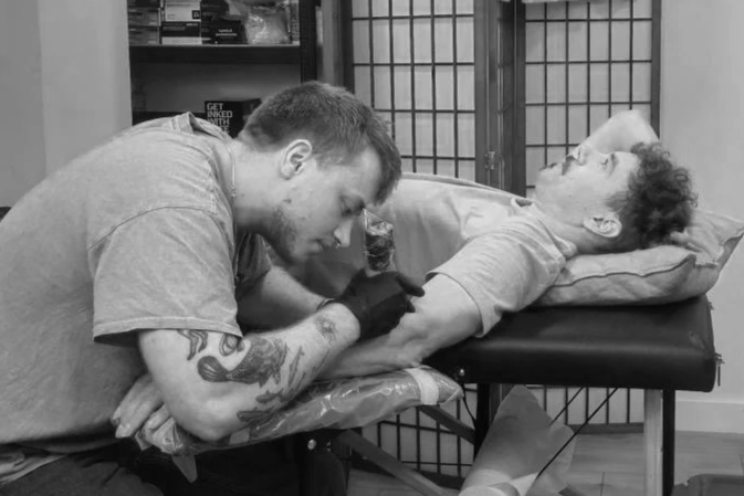 A tattooed man giving a tattoo to  man lying on a medical table in a room with a folding privacy screen.