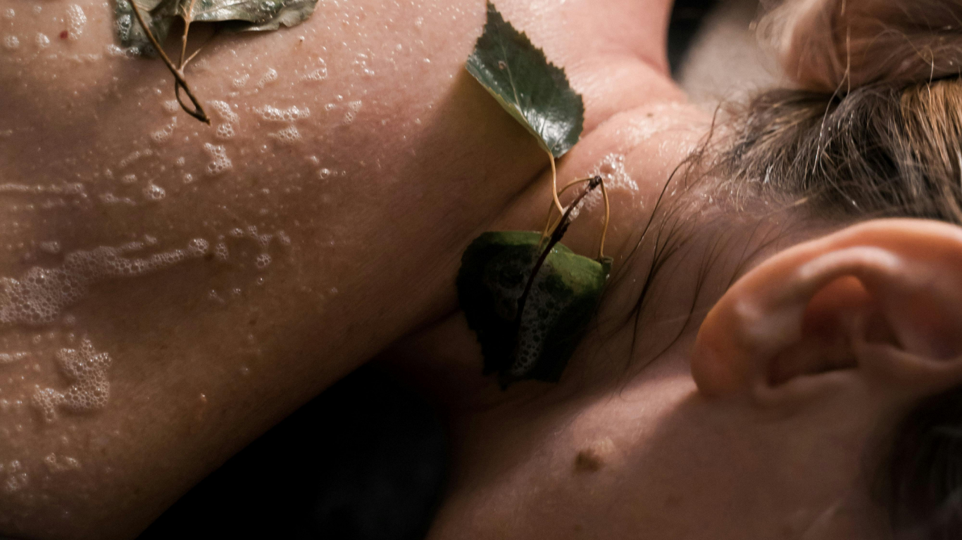 Close-up of a person receiving a facial treatment with leaves and foam on their face, lying down with their eyes closed.