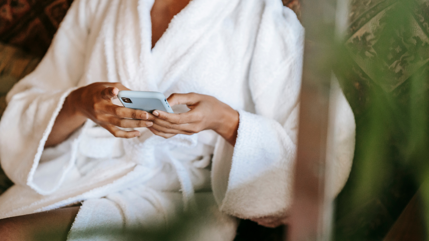 A person in a white bathrobe sitting indoors, holding and looking at a smartphone.