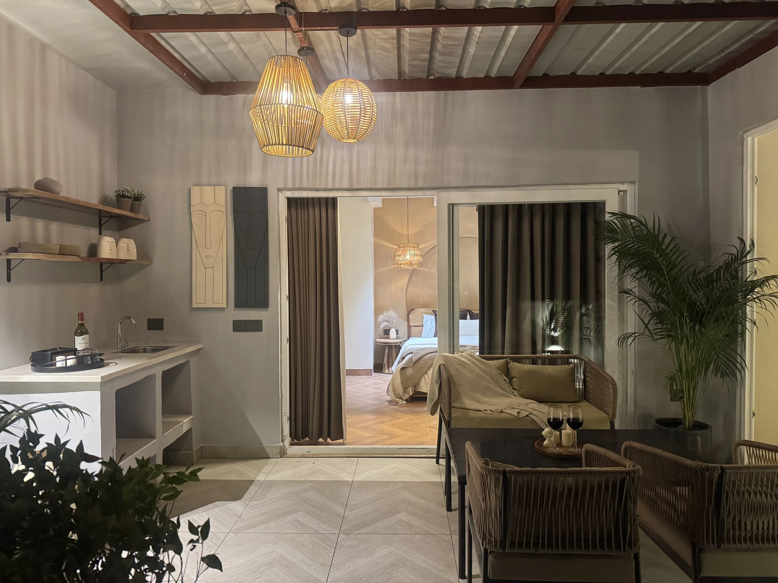 Living room with rattan chairs, a black dining table with two glasses of red wine, a beige sofa with a blanket, potted plant, open sliding door to bedroom, hanging pendant lights, shelves with decor on wall, small kitchenette with sink and bottles, and a bedroom visible through sliding doors with bed and ceiling chandelier.