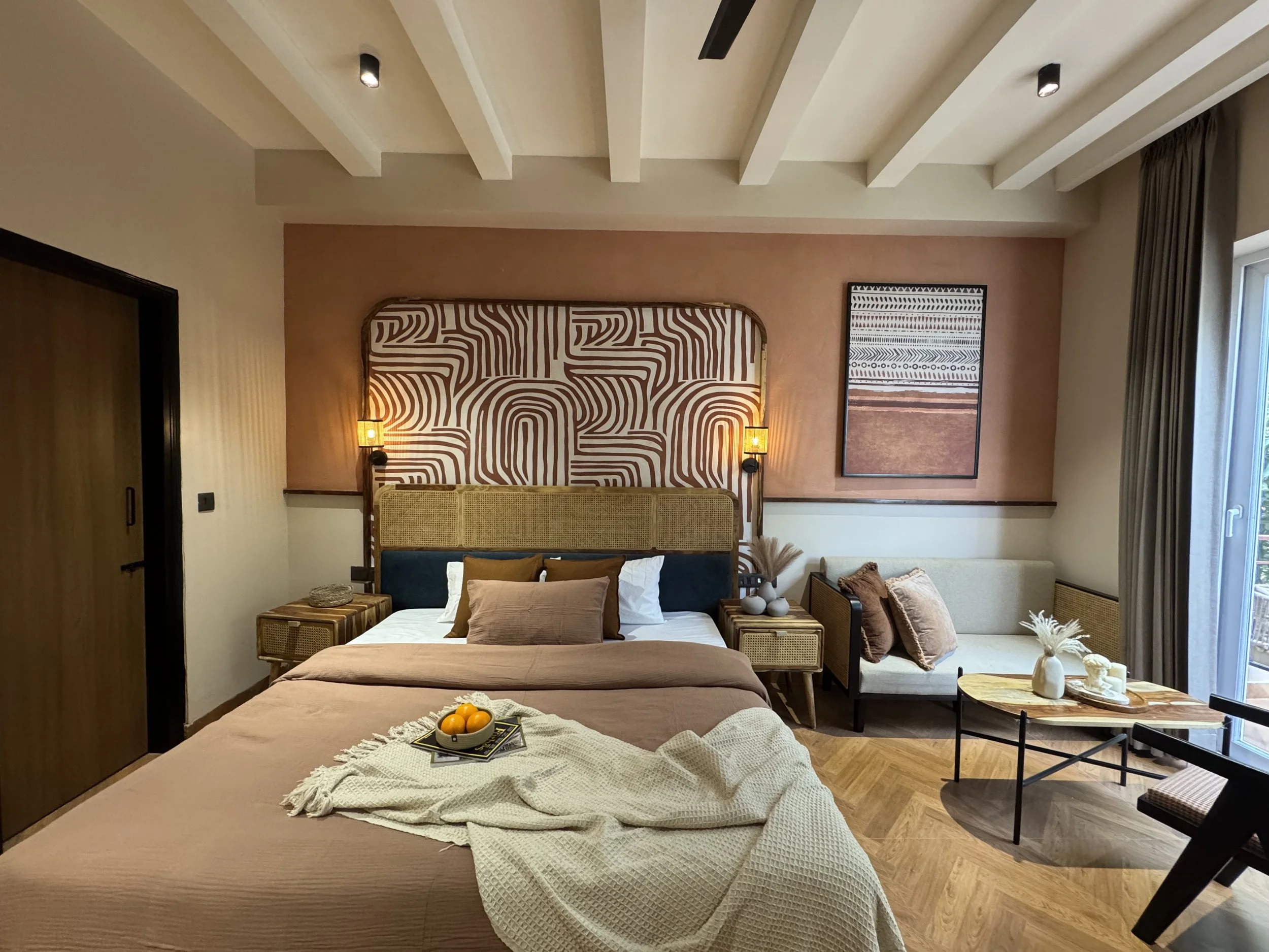 A modern bedroom with a large bed, decorative pillows, and a tray of oranges, accented with warm peach and beige tones, artwork, and natural light from a window.