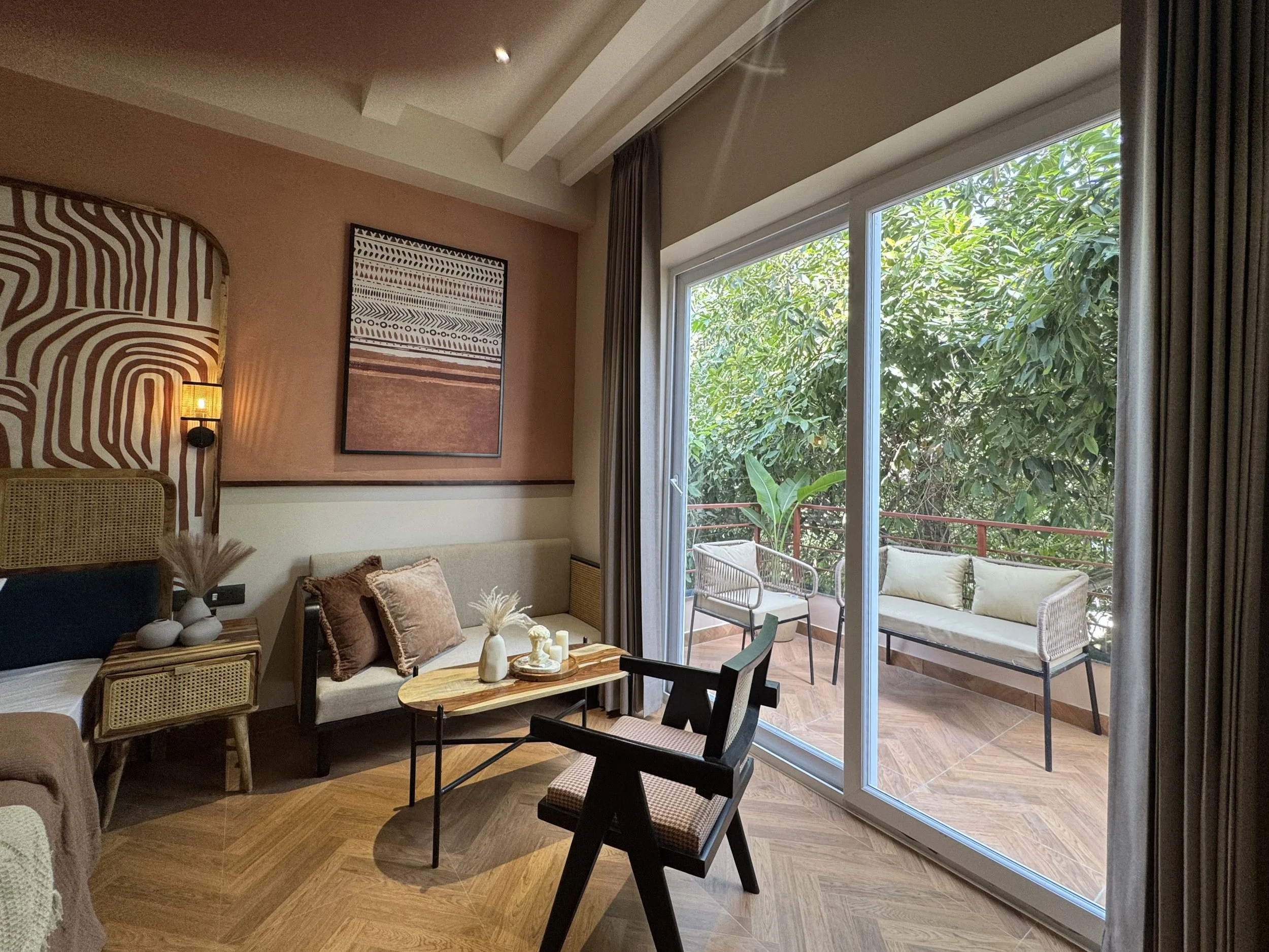 Interior of a cozy living room with a sliding glass door leading to a small balcony with outdoor seating, lush green trees outside, a beige sofa with pillows, a wooden coffee table with decorative items, a black armchair, and abstract wall art.