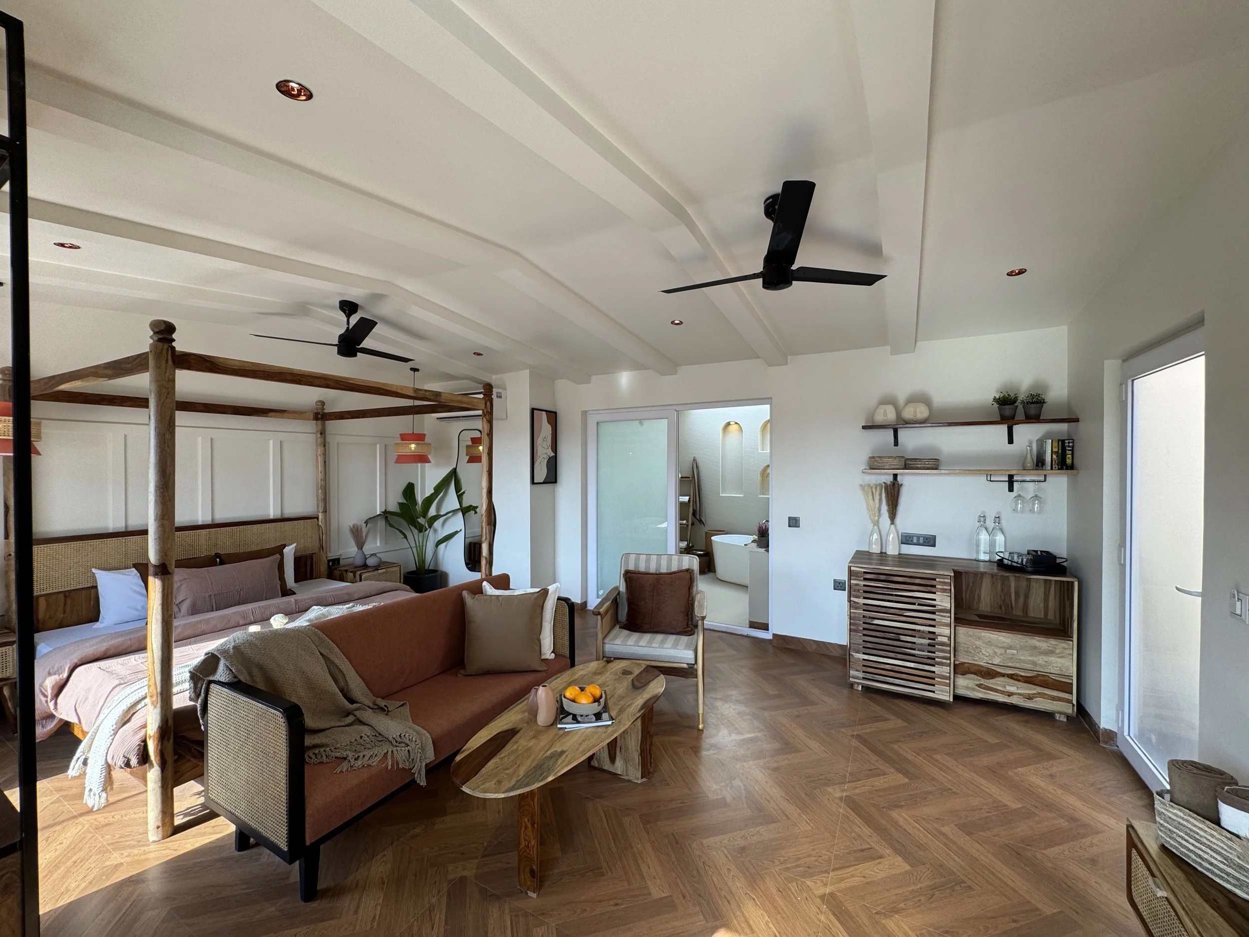 A spacious living area with a wooden four-poster bed, a comfortable sofa, a wooden coffee table with oranges, and a small cabinet with decorative items, in a modern and cozy style.