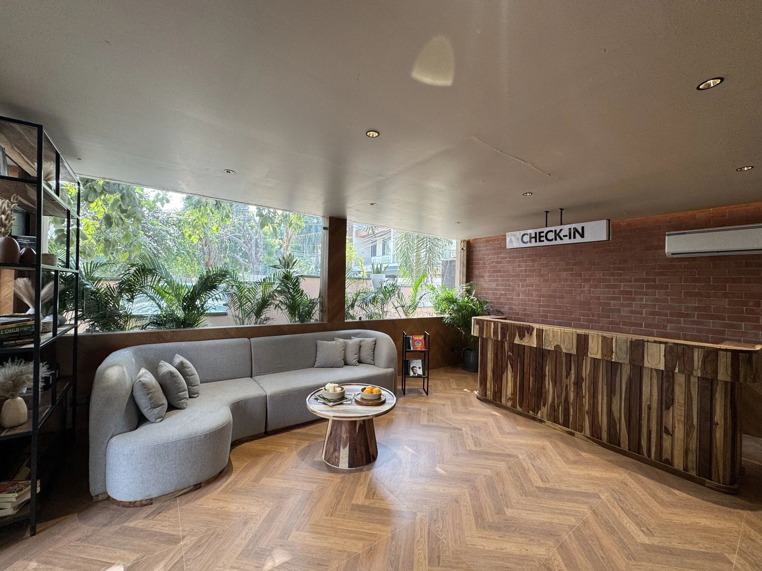 Hotel lobby reception area with a check-in sign, beige curved sofa, wooden coffee table with a bowl of snacks, background with large windows, indoor plants, brick wall, wooden reception desk, and bookshelf with decorative items.