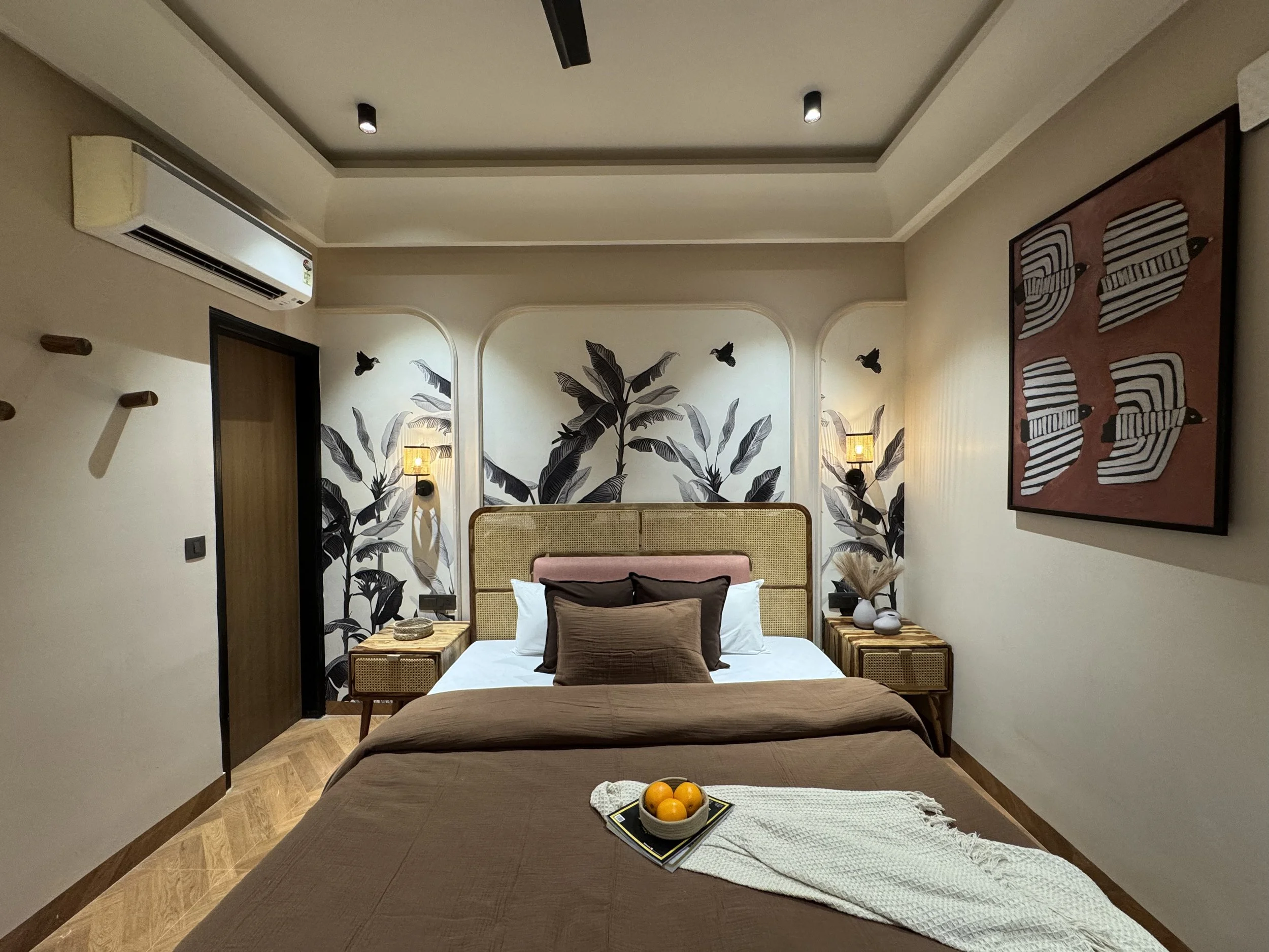 Modern bedroom with a bed, artwork, wall-mounted lamps, and decorative textiles, featuring tropical leaf and animal patterns.