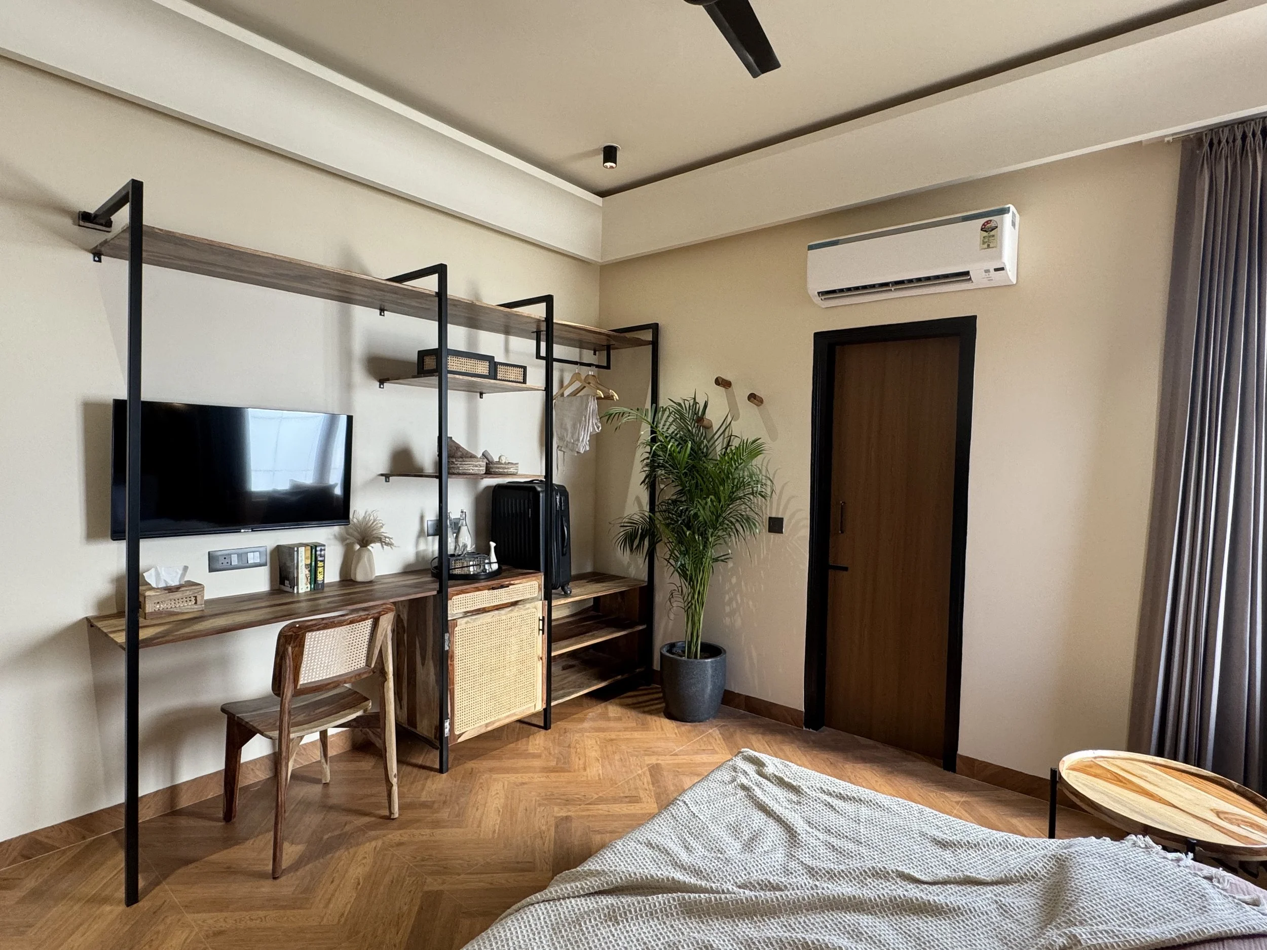 Modern bedroom corner with a wall-mounted TV, a wooden desk with a woven chair, shelves, a suitcase, and a large potted plant next to a wooden door; beige walls, wooden flooring, and curtains on the window.