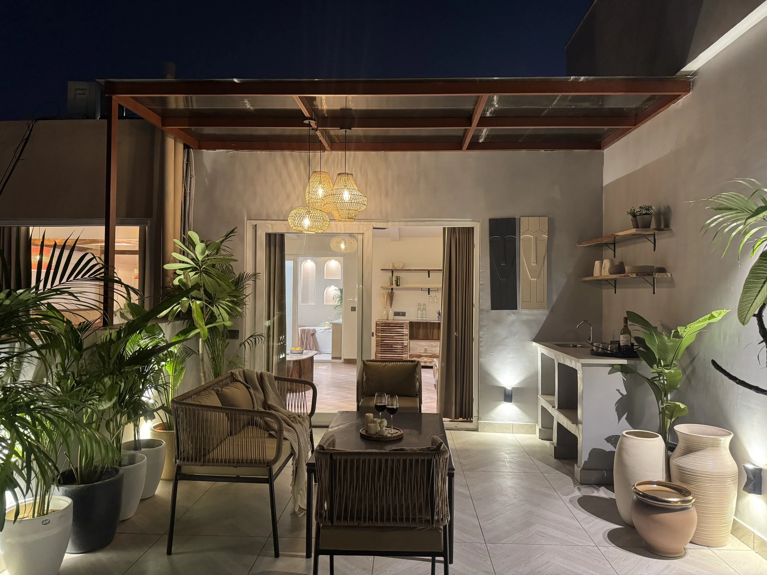 Nighttime view of a cozy outdoor balcony with seating, plants, decorative lighting, and a small kitchenette area.