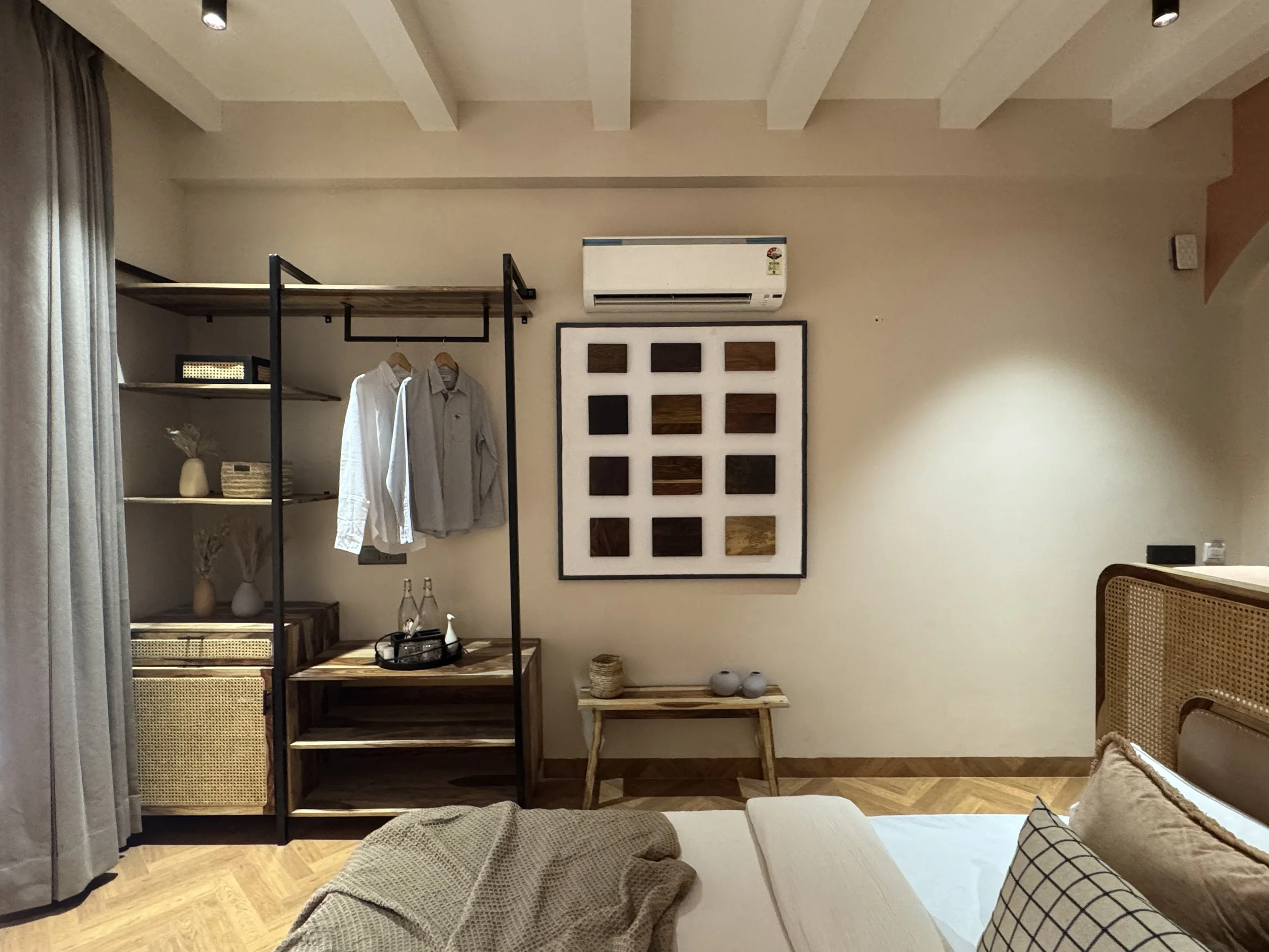 A bedroom with a bed, pillows, a clothing rack with shirts, a wall-mounted air conditioner, a framed artwork with squares of wood, a small wooden table, and side tables with vases and bottles.