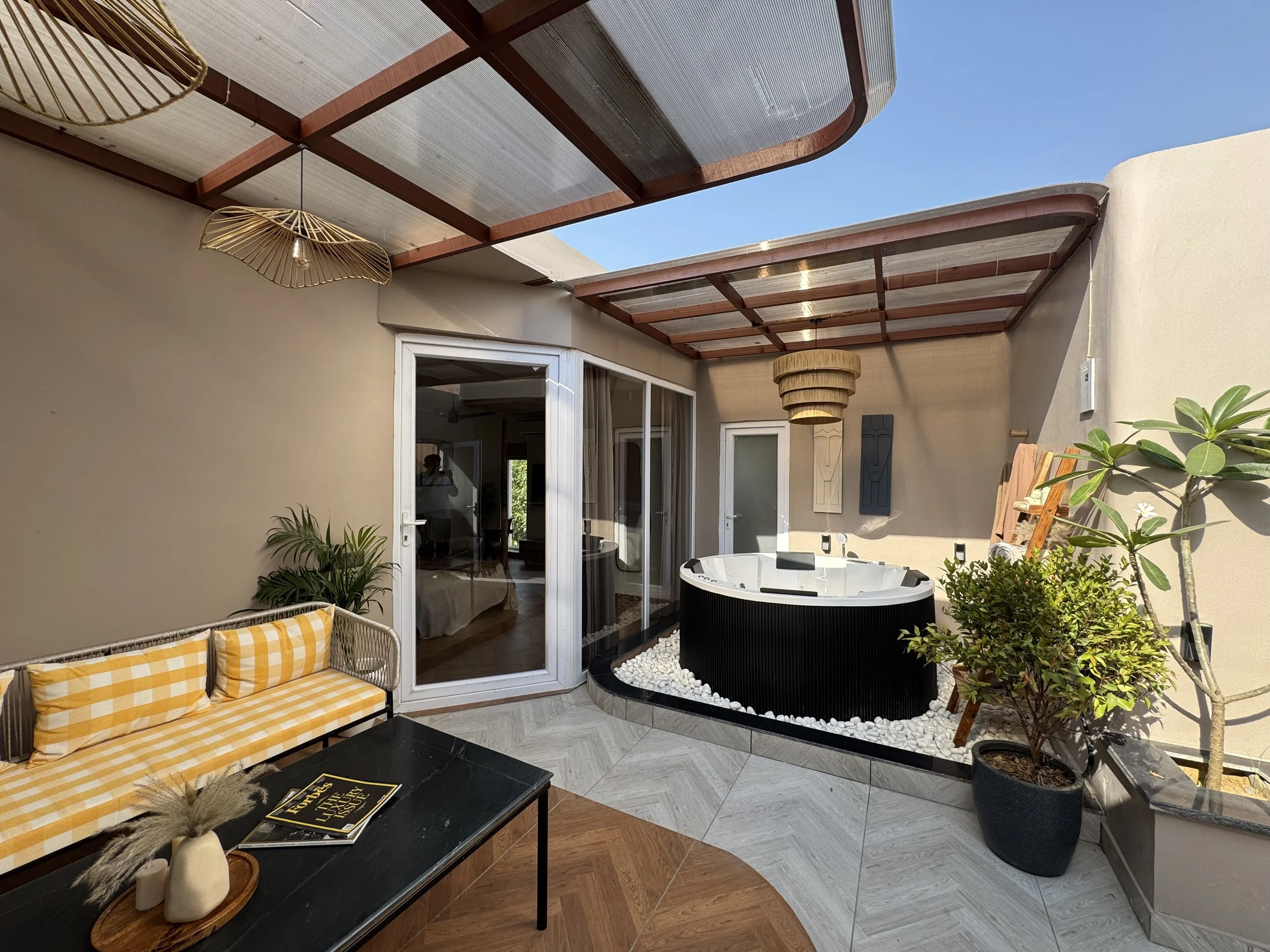 Outdoor patio area with a hot tub, yellow checkered bench, and potted plants, under a partially transparent roof, with a sliding glass door leading indoors.