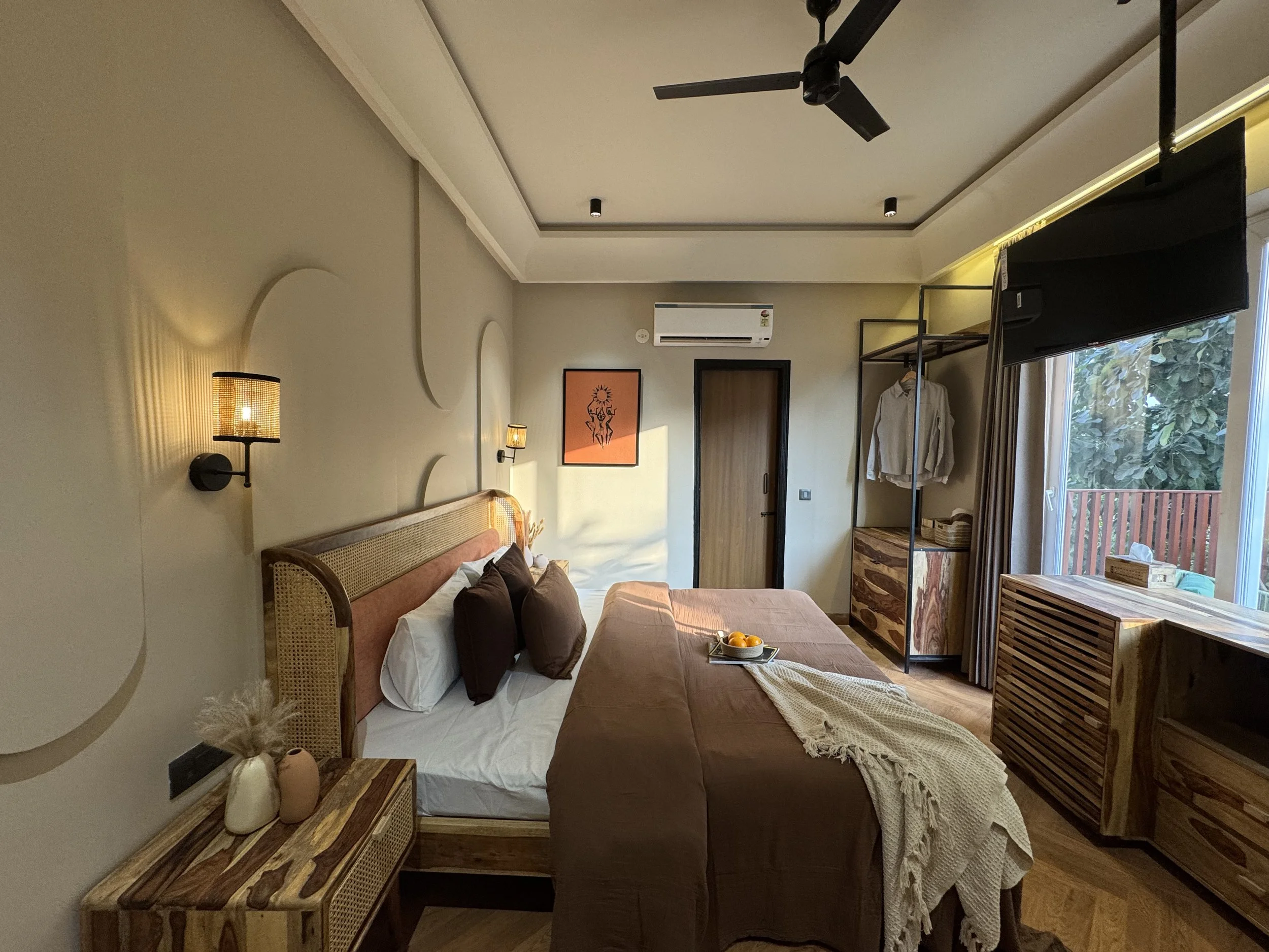 Modern bedroom with a bed, brown bedding, pillows, wall-mounted lamps, a dresser, and a large window with curtains and greenery outside.