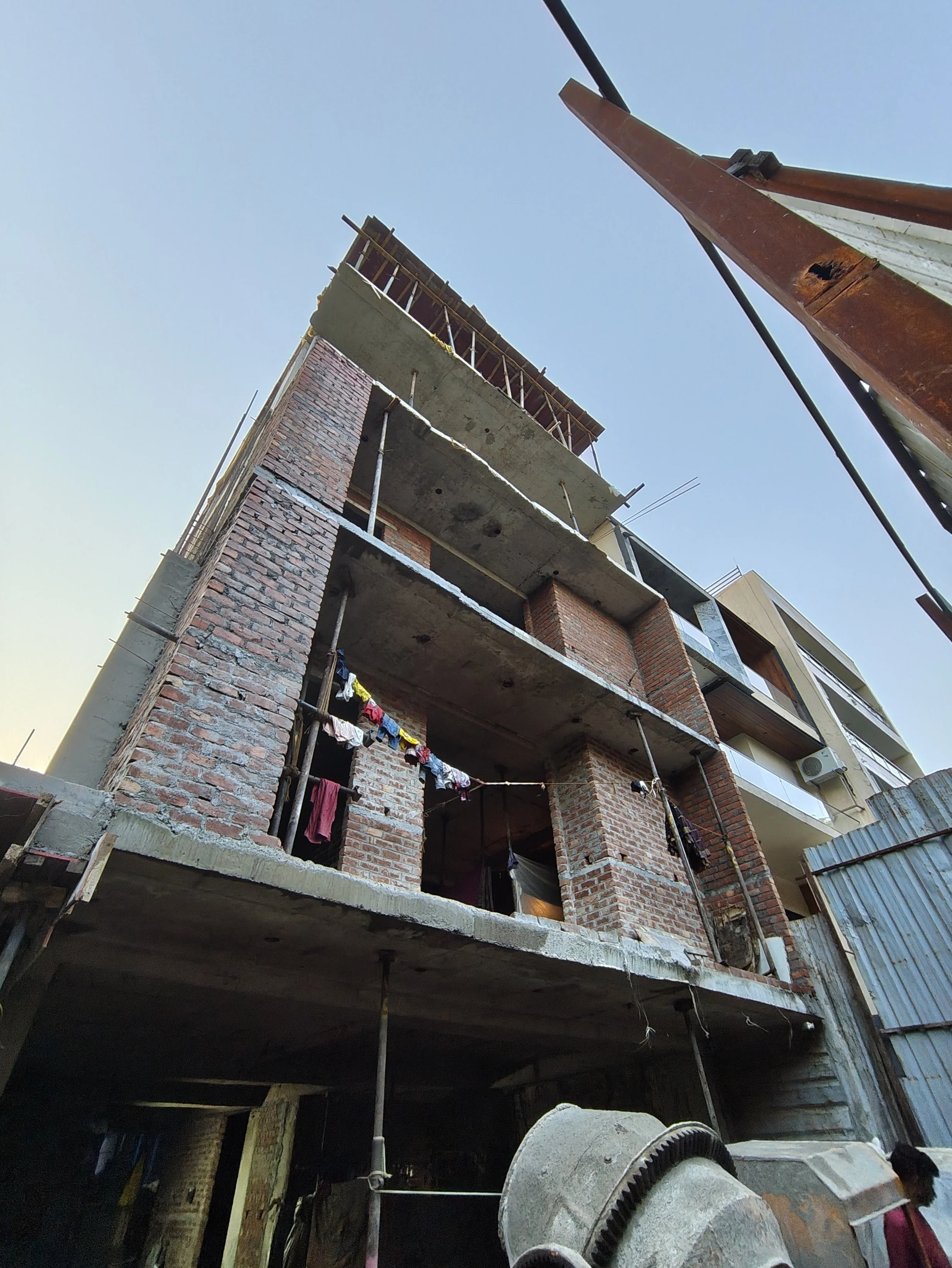 A multi-story building under construction with exposed brick walls and concrete floors, laundry hanging outside on one level, and construction equipment at the base.