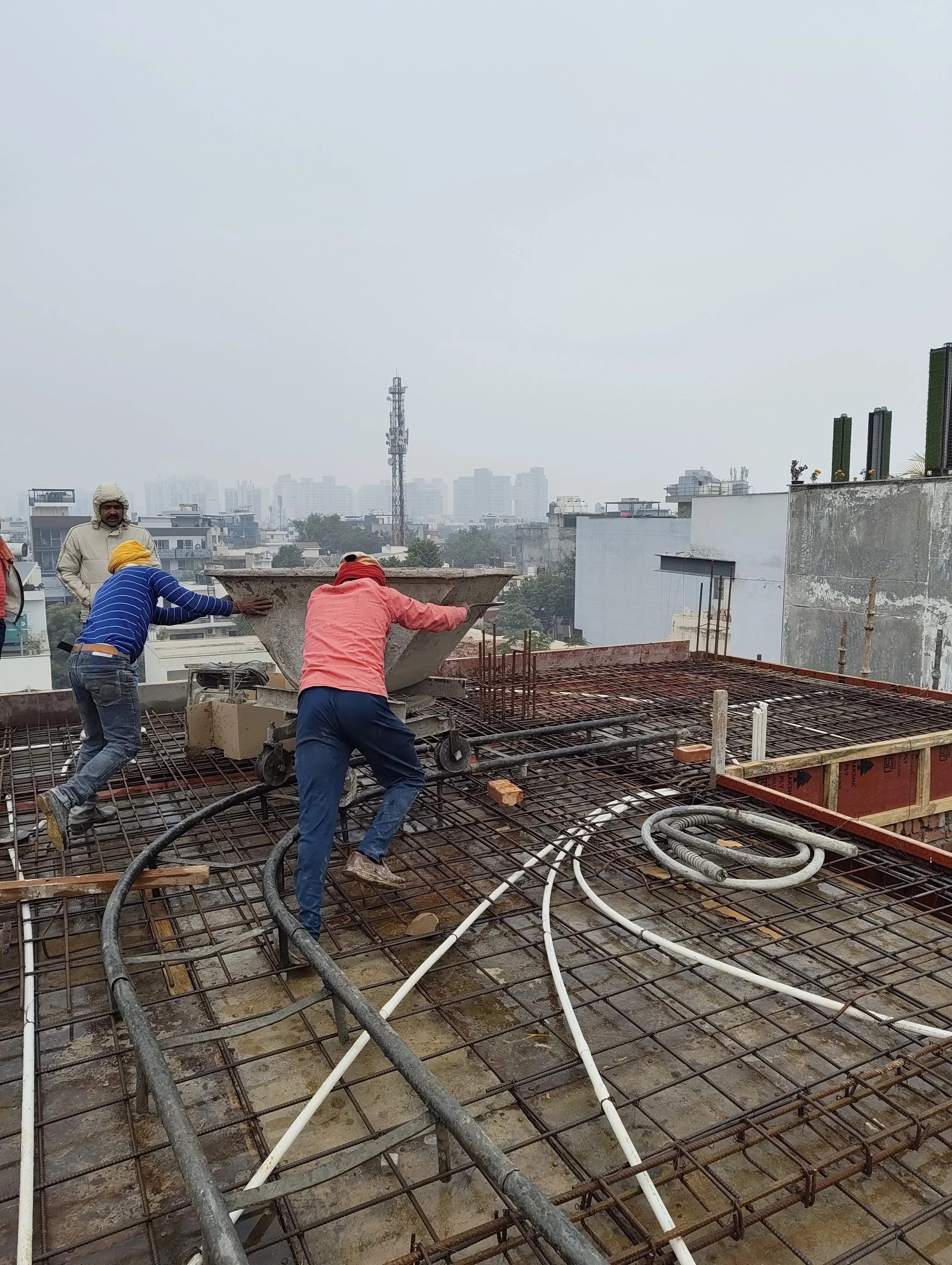 Construction workers working on a building rooftop, pouring concrete over steel rebar with a city skyline in the background.