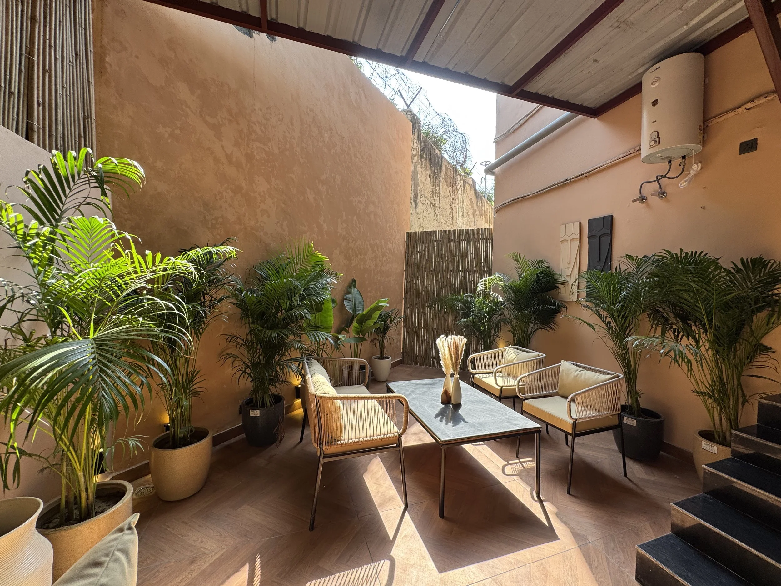 Sunlit outdoor patio with beige walls, lush potted green plants, a black table, and four white cushioned chairs.